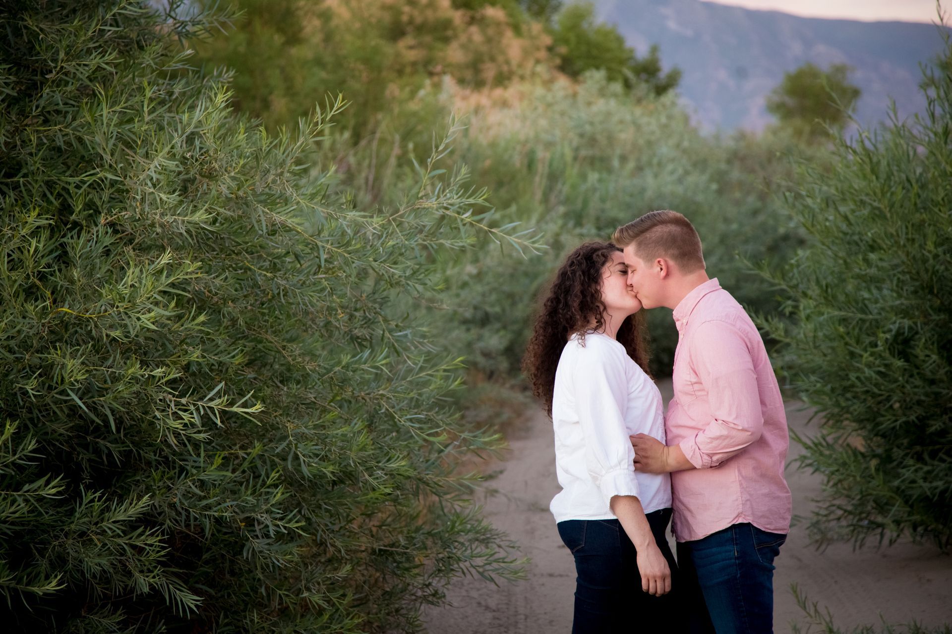 A couple standing on a dirt path between green trees, leaning in toward each other to kiss in an outdoor setting.
