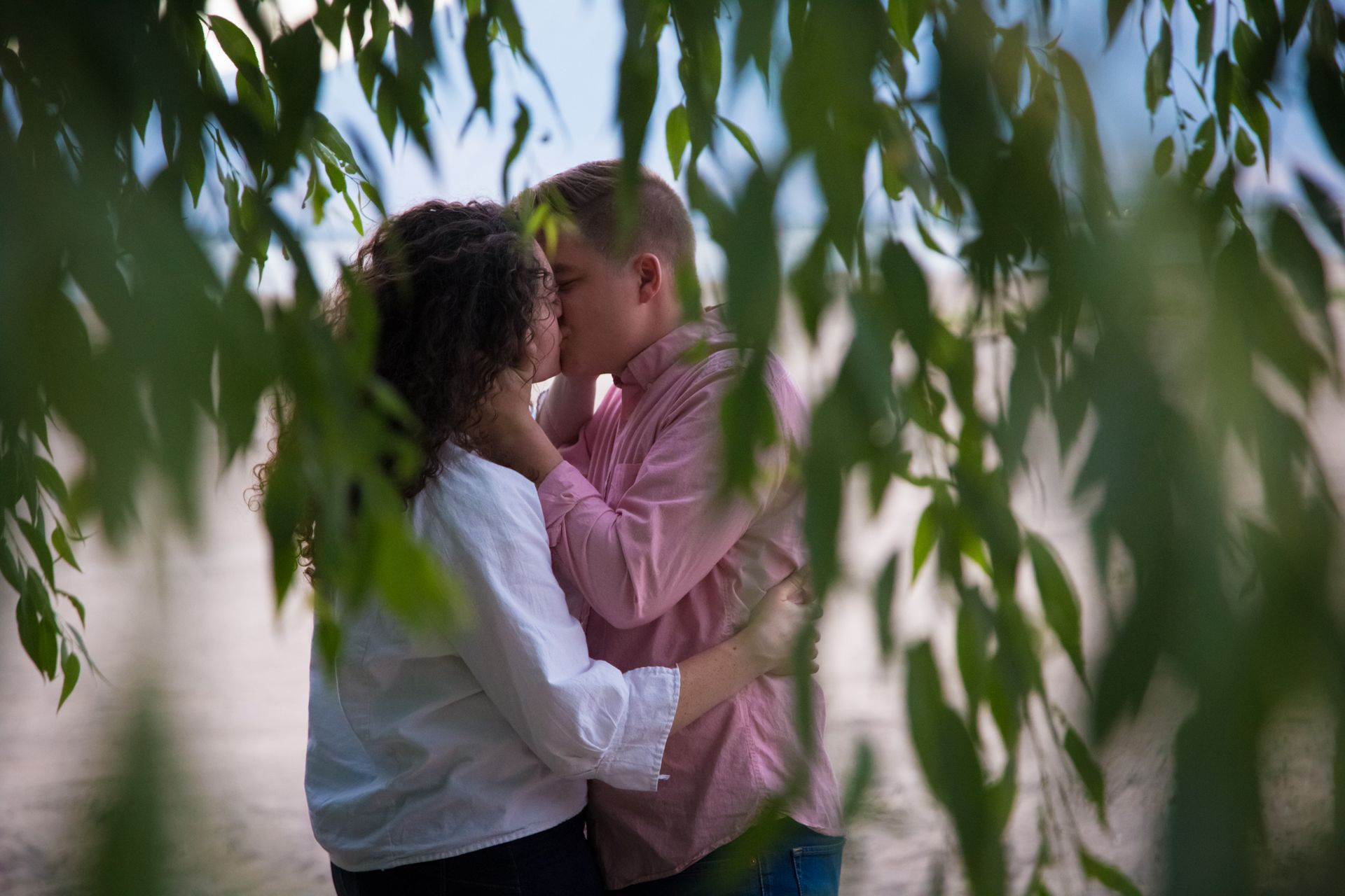 A couple kisses while standing together outdoors, partially framed by hanging willow tree branches.