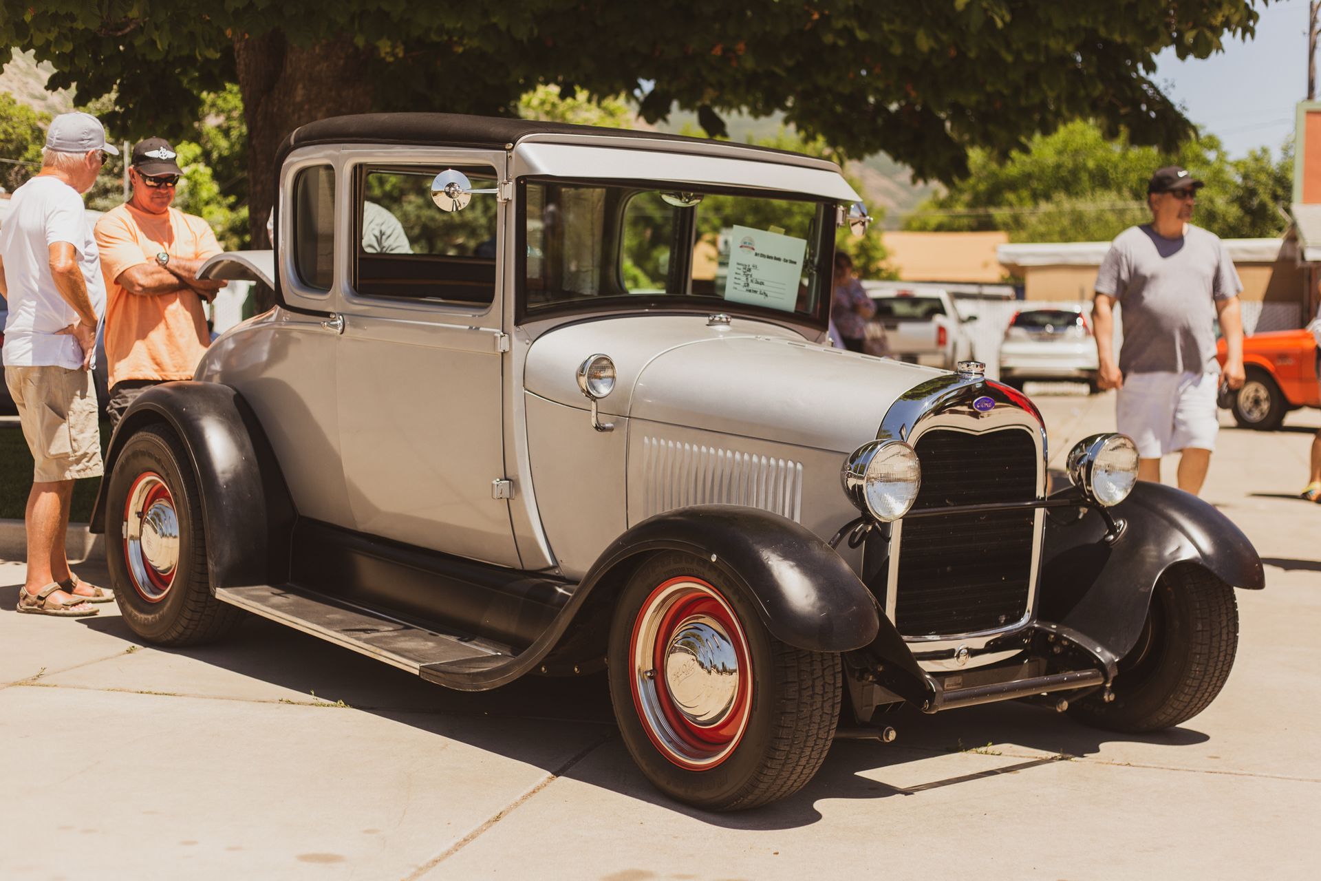 A classic light grey vintage coupe with black fenders on display at an outdoor car show with several people standing by.