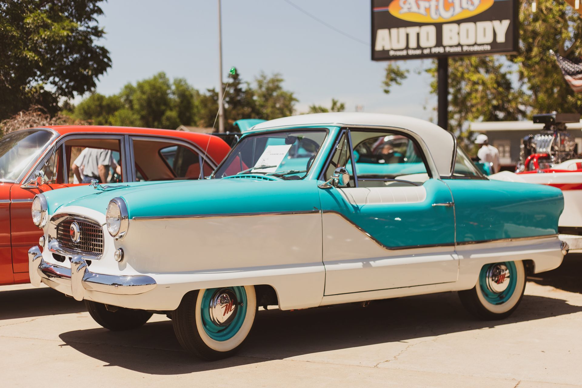 A two-tone teal and white vintage Nash Metropolitan convertible parked at an outdoor car show.
