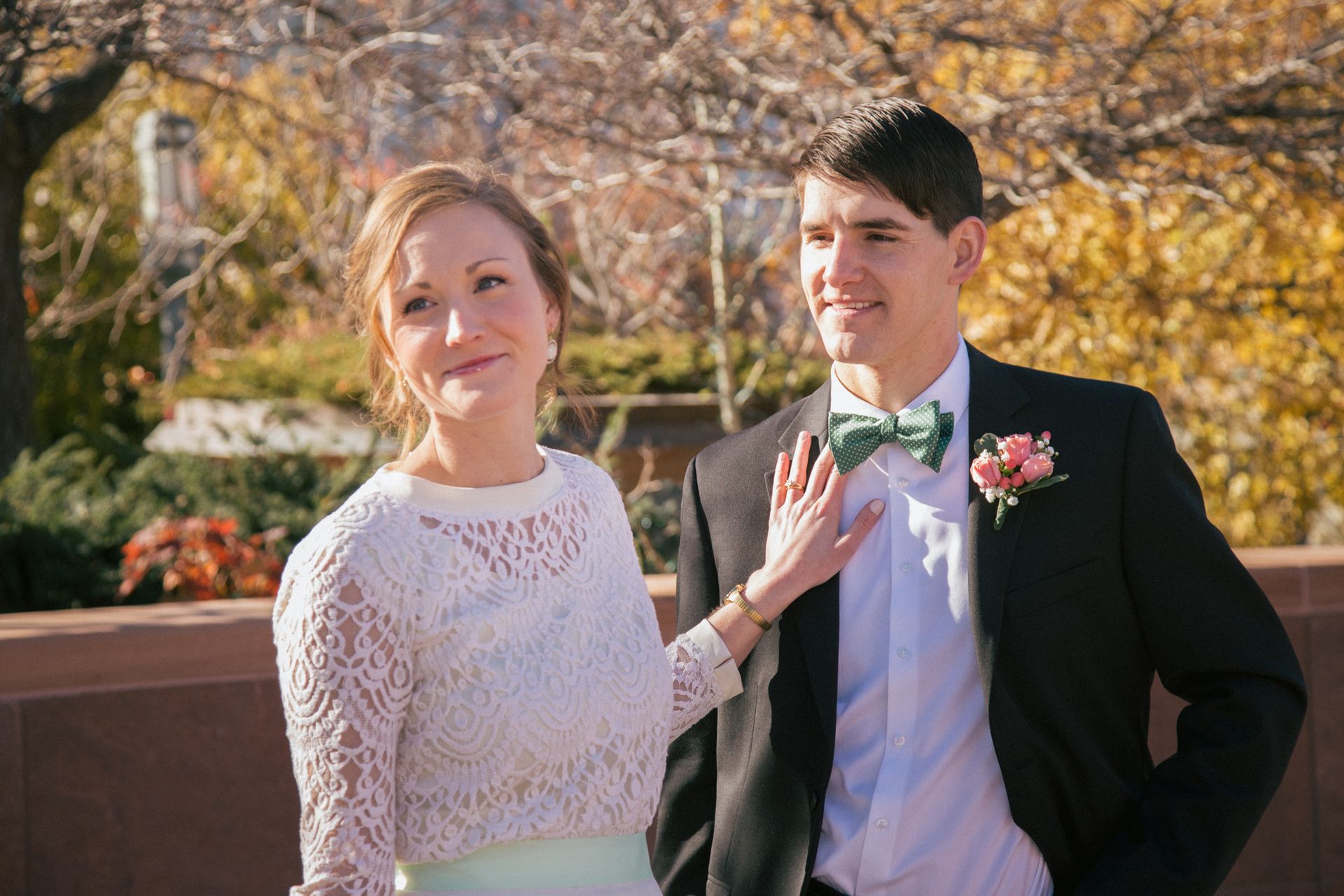 A couple in wedding attire stands outdoors, with the person on the left touching the other's teal bowtie.