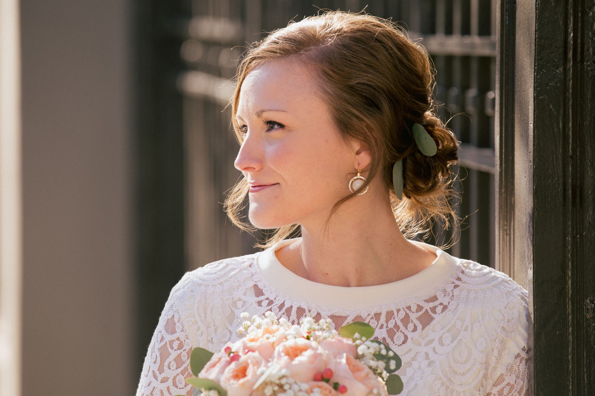 A person in a white lace dress with an updo hairstyle holding a bouquet of pink roses, looking to the side.