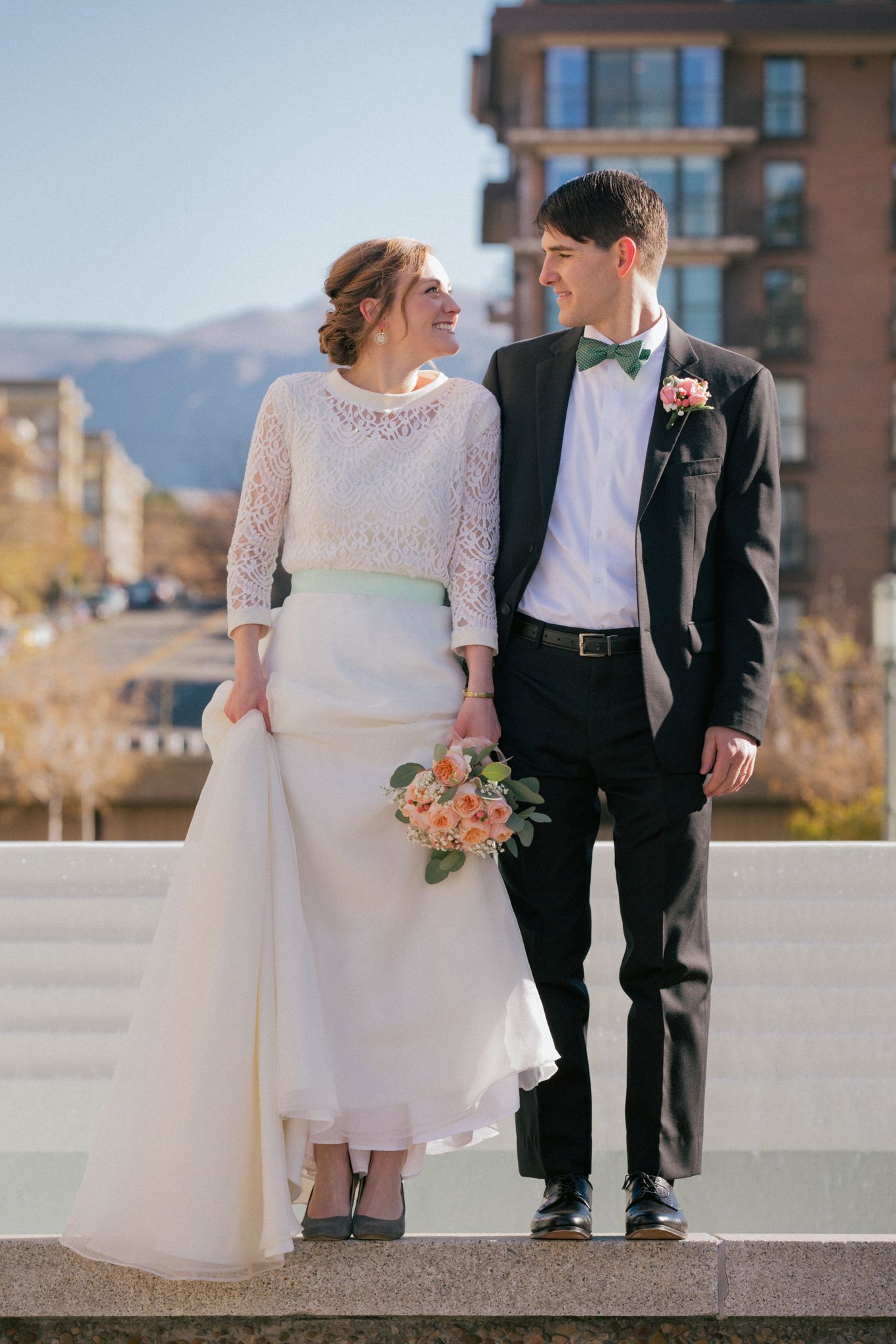 A smiling couple in wedding attire holding a bouquet, standing outdoors with a city and mountain backdrop.