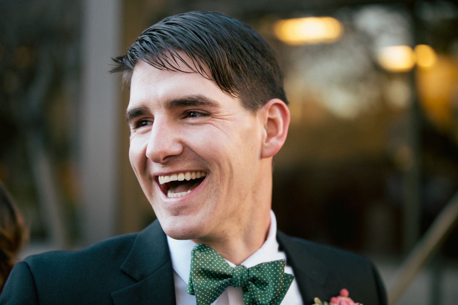 A person in a black suit and green patterned bow tie laughing, shown from the chest up.