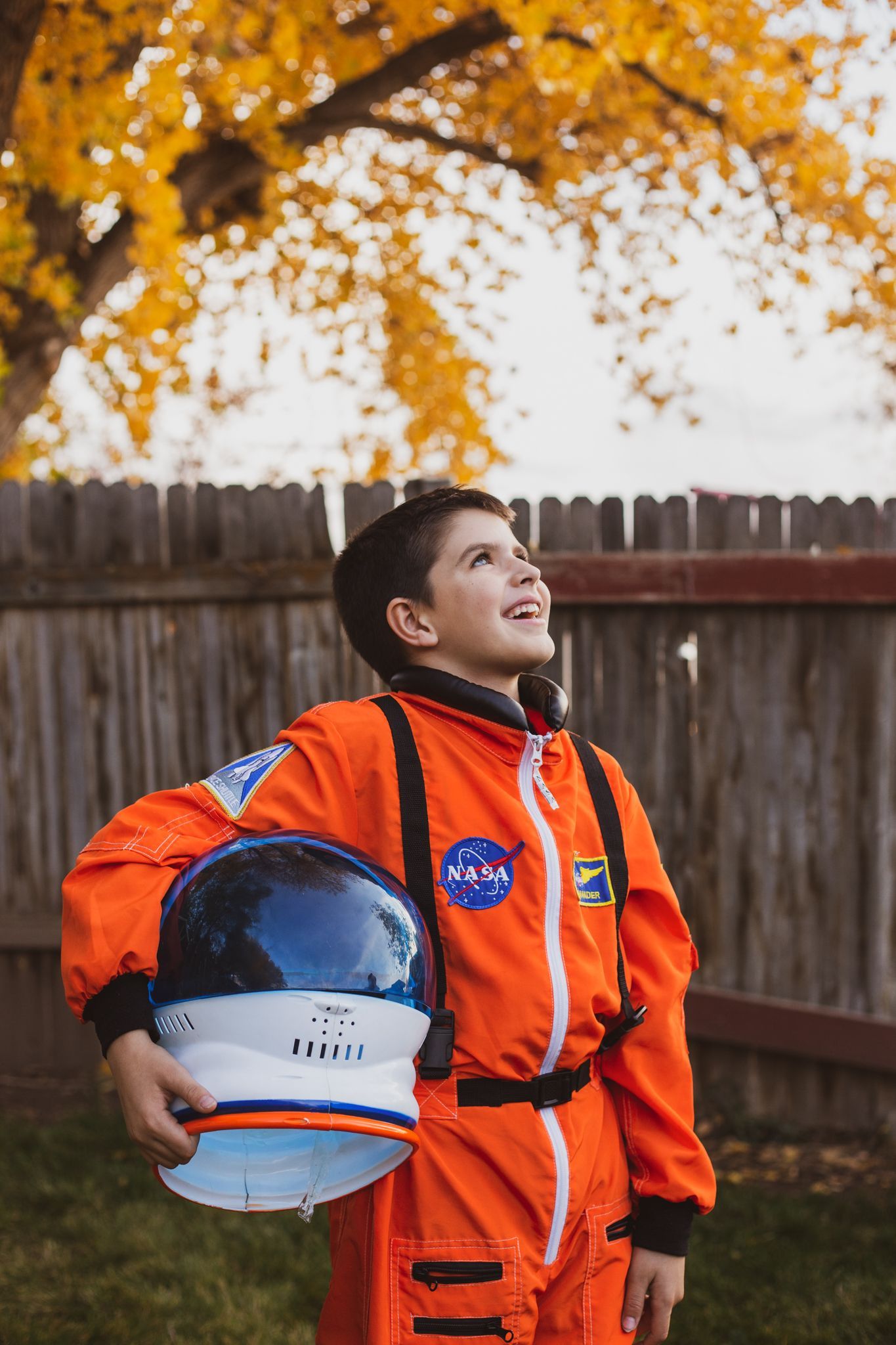 A child in an orange astronaut suit holds a helmet, looking upward in a backyard with autumn trees.