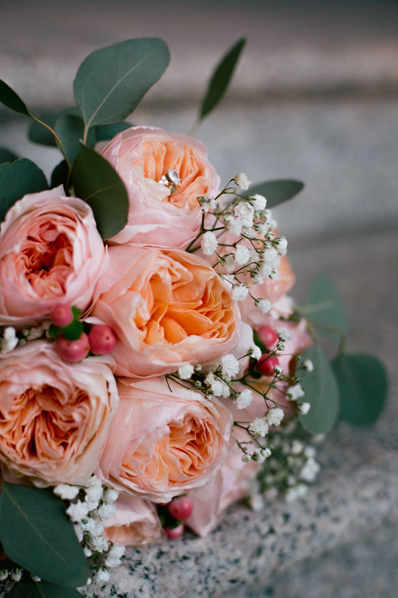 A wedding bouquet of soft peach and pink garden roses, white baby's breath, and eucalyptus leaves on a stone surface.