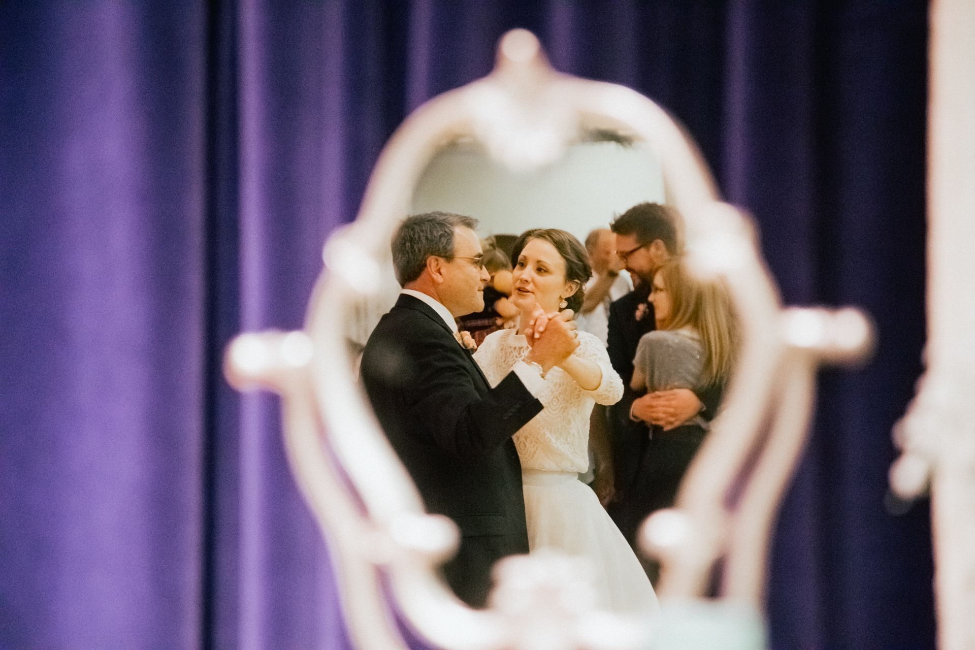 A couple dances in the reflection of an ornate mirror, set against a backdrop of draped purple curtains.
