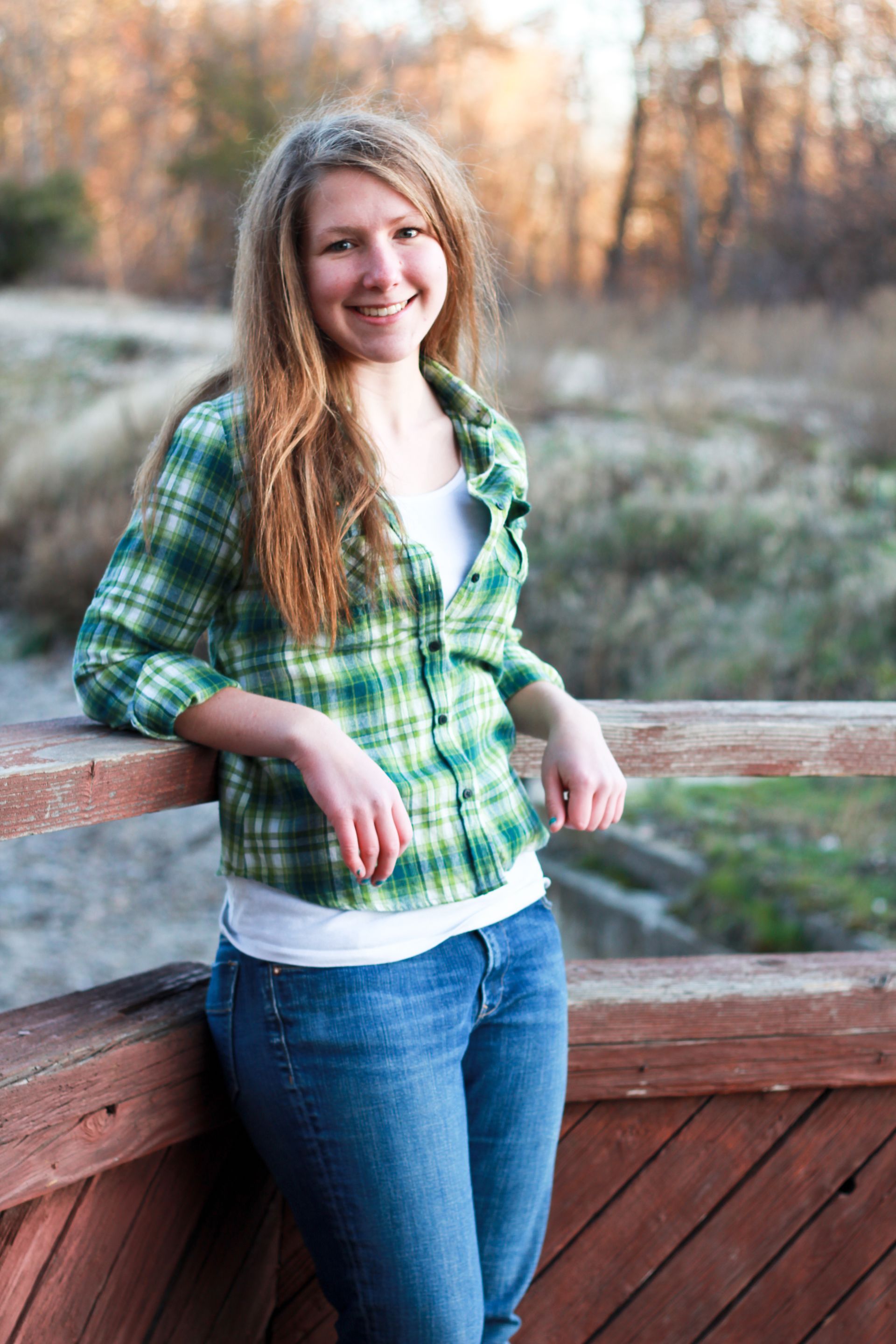A smiling person with long hair wearing a green plaid shirt and jeans, leaning on a wooden railing outdoors.