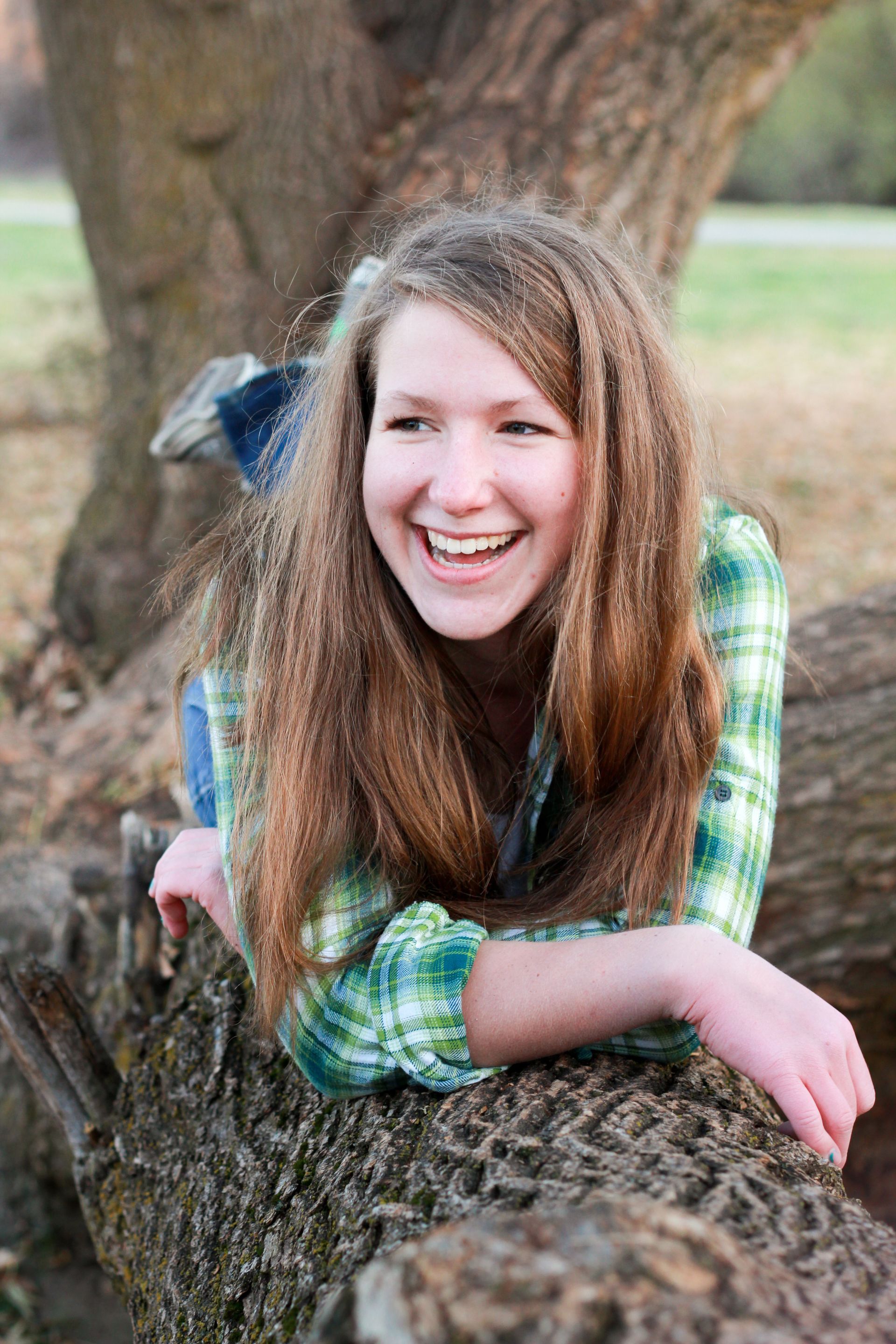 A person with long, wavy brown hair wearing a green plaid shirt, smiling while lying across a fallen tree trunk outdoors.