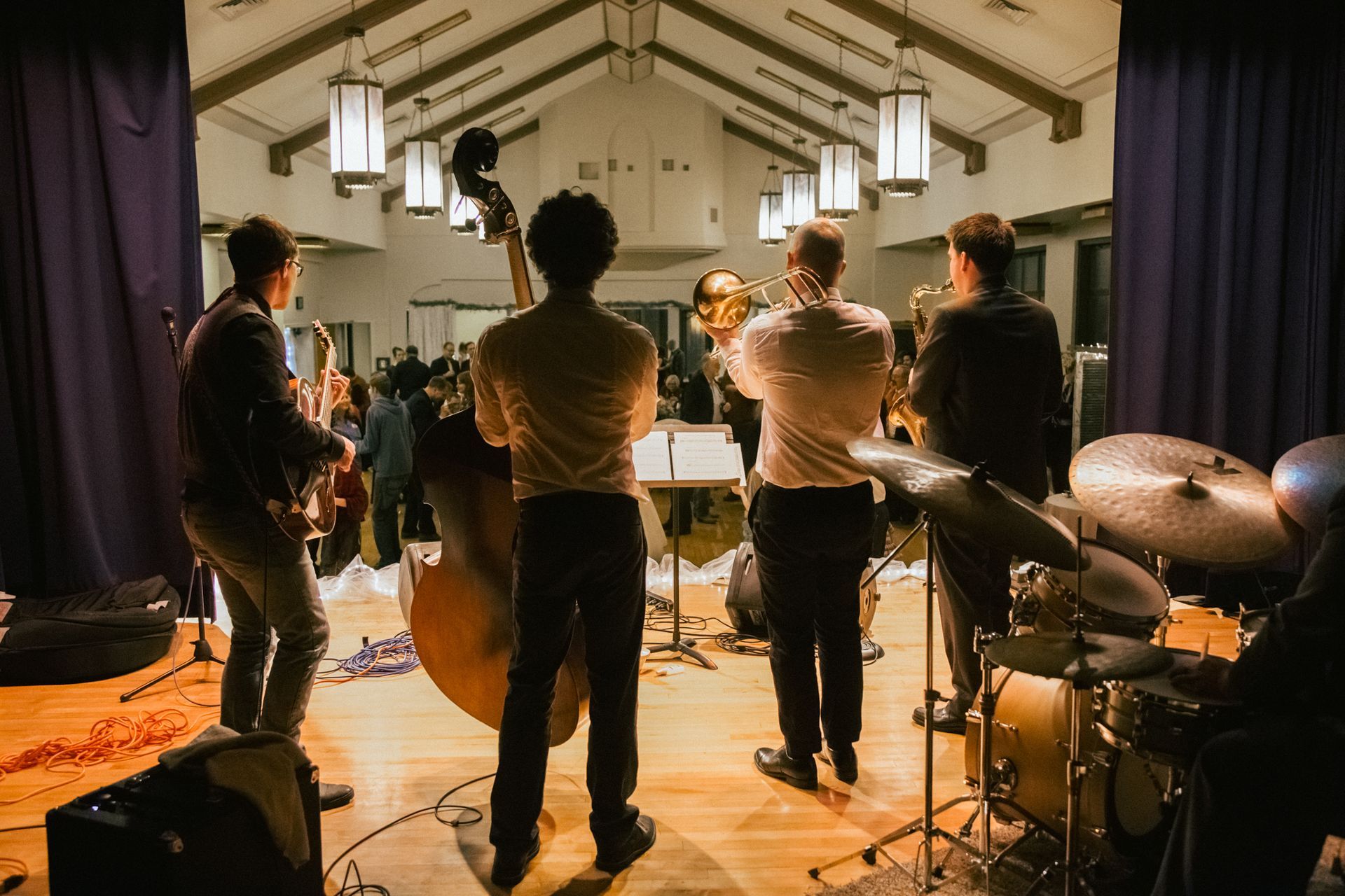 A small band plays on a wooden stage in a dimly lit hall, featuring a double bass, trumpet, guitar, and drum set.