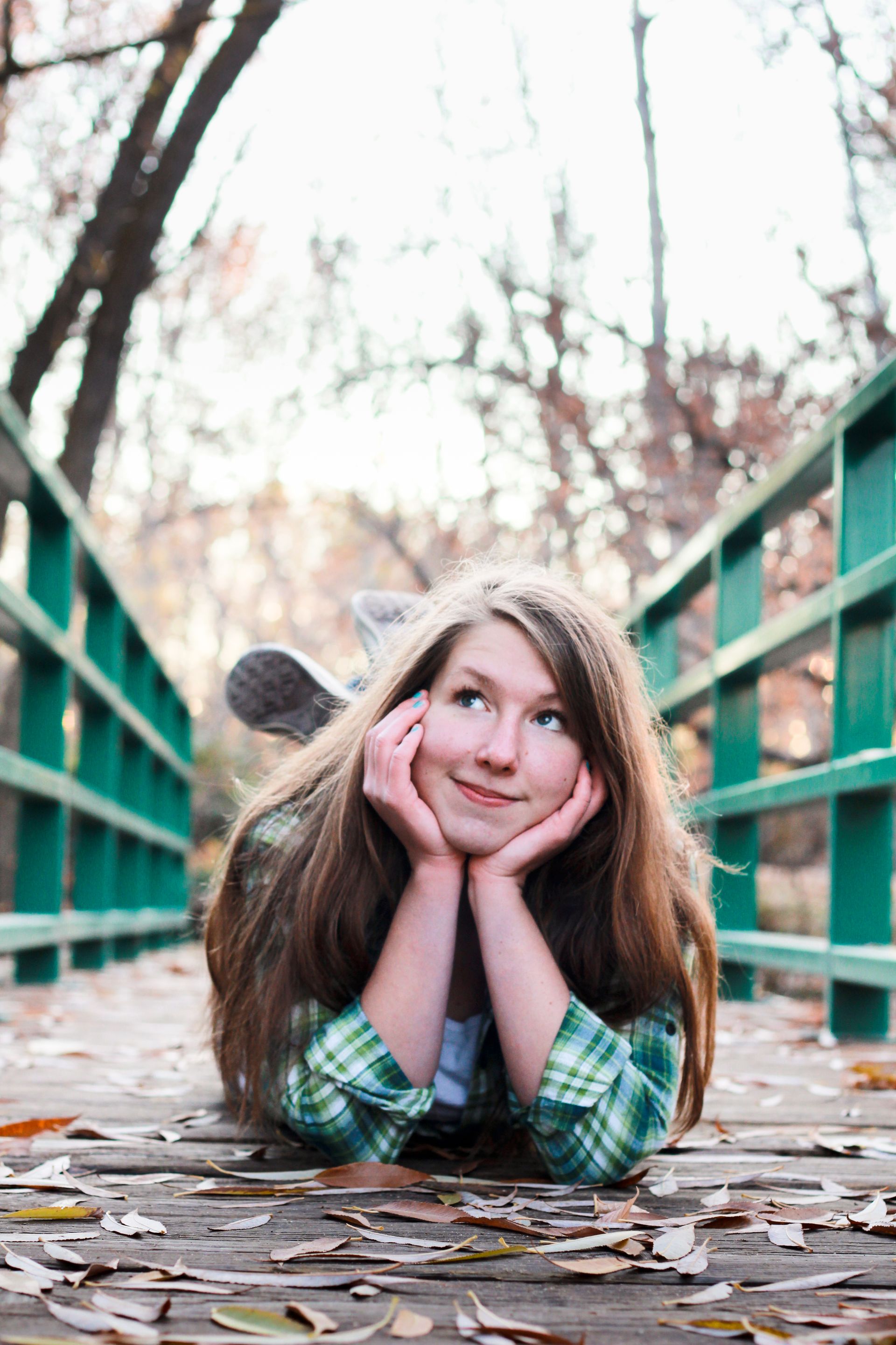 A person lying on a wooden bridge strewn with autumn leaves, resting their chin on their hands and looking upward.