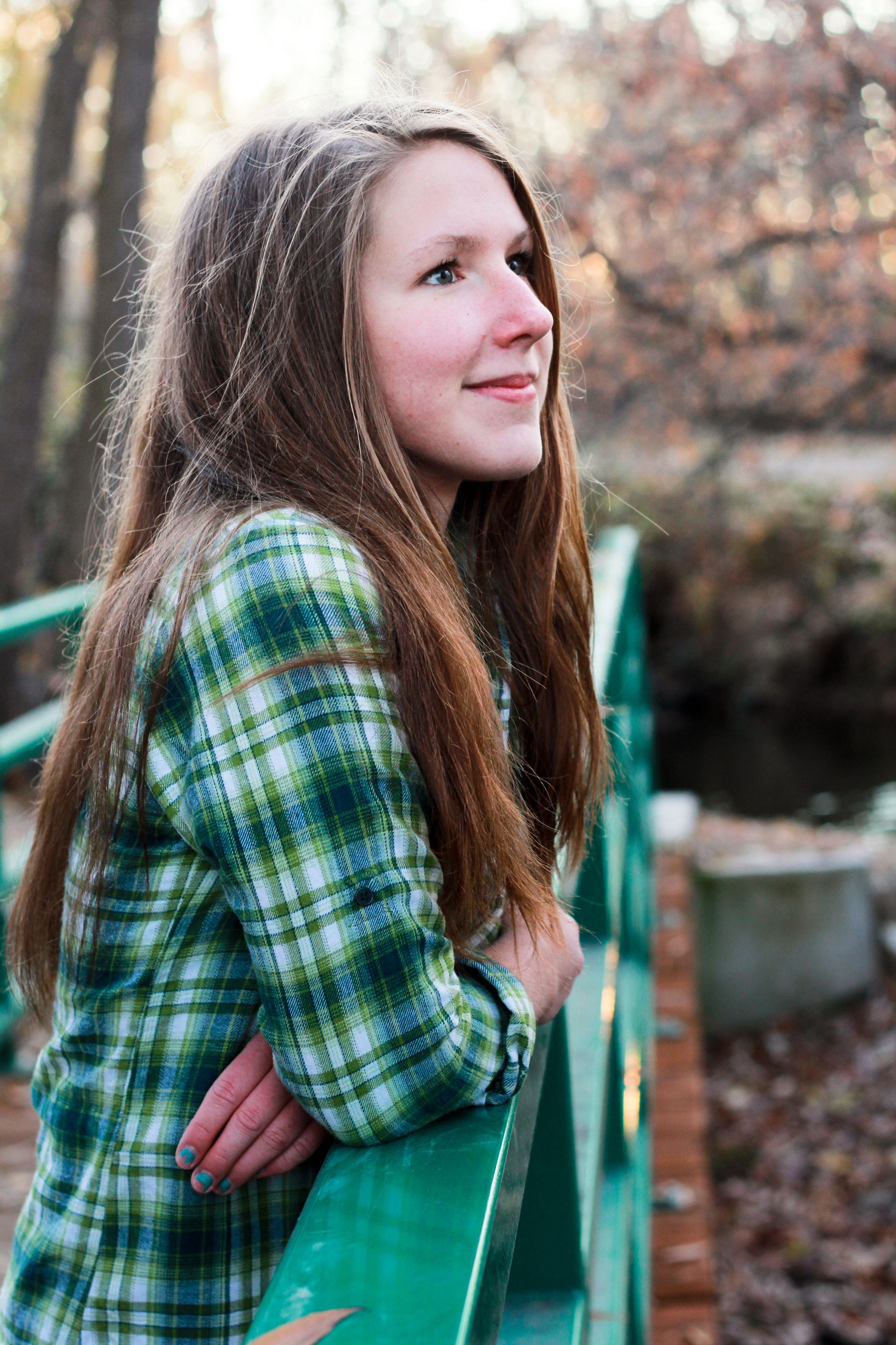 A person with long hair wears a green plaid shirt while leaning on a green bridge railing in a wooded outdoor setting.