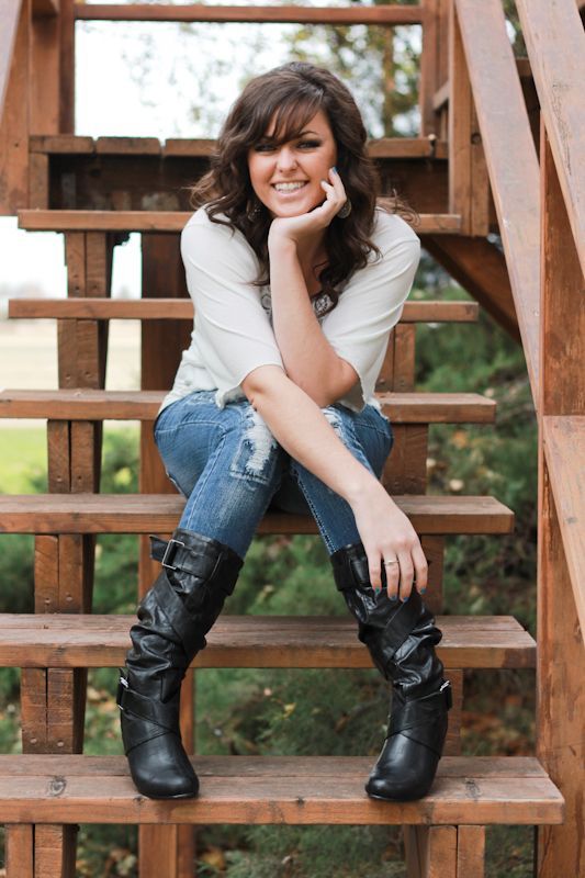 A person with dark hair sits on wooden stairs, wearing a white shirt, blue jeans, and black boots, smiling at the camera.