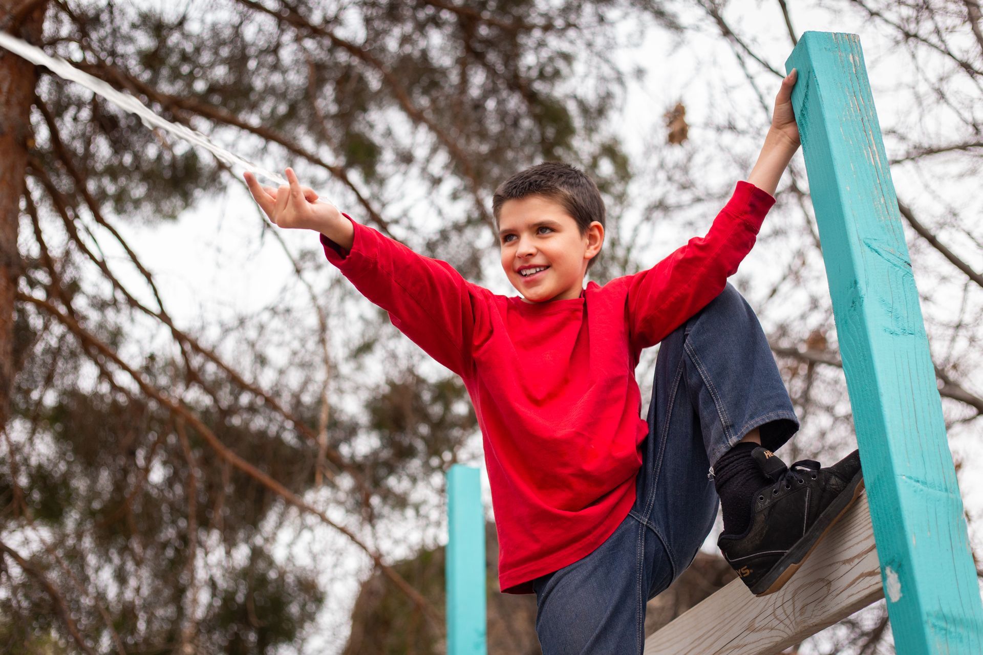 A smiling child in a red long-sleeved shirt climbs a blue play structure outdoors with trees in the background.