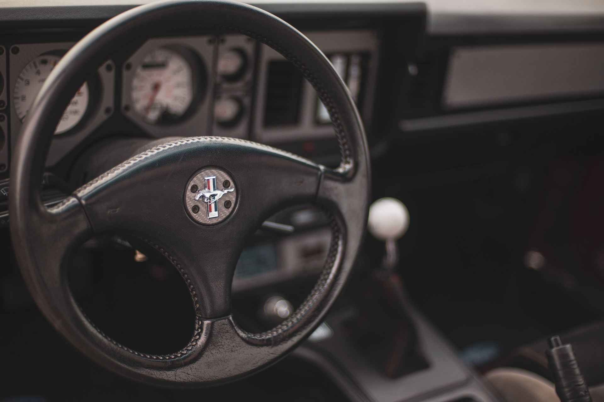Interior view of a black sports car dashboard and steering wheel with a circular logo, featuring a white gear shift knob.