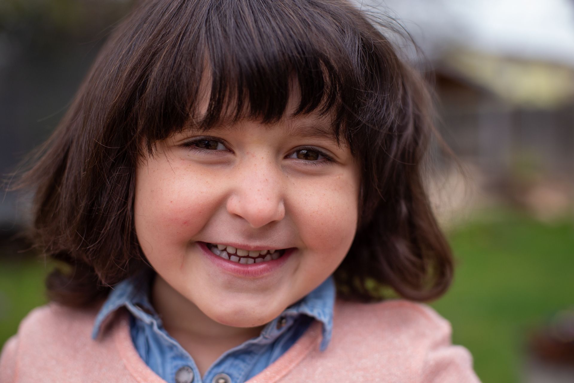 A smiling child with dark, shoulder-length hair, wearing a light pink sweater over a blue collared shirt.
