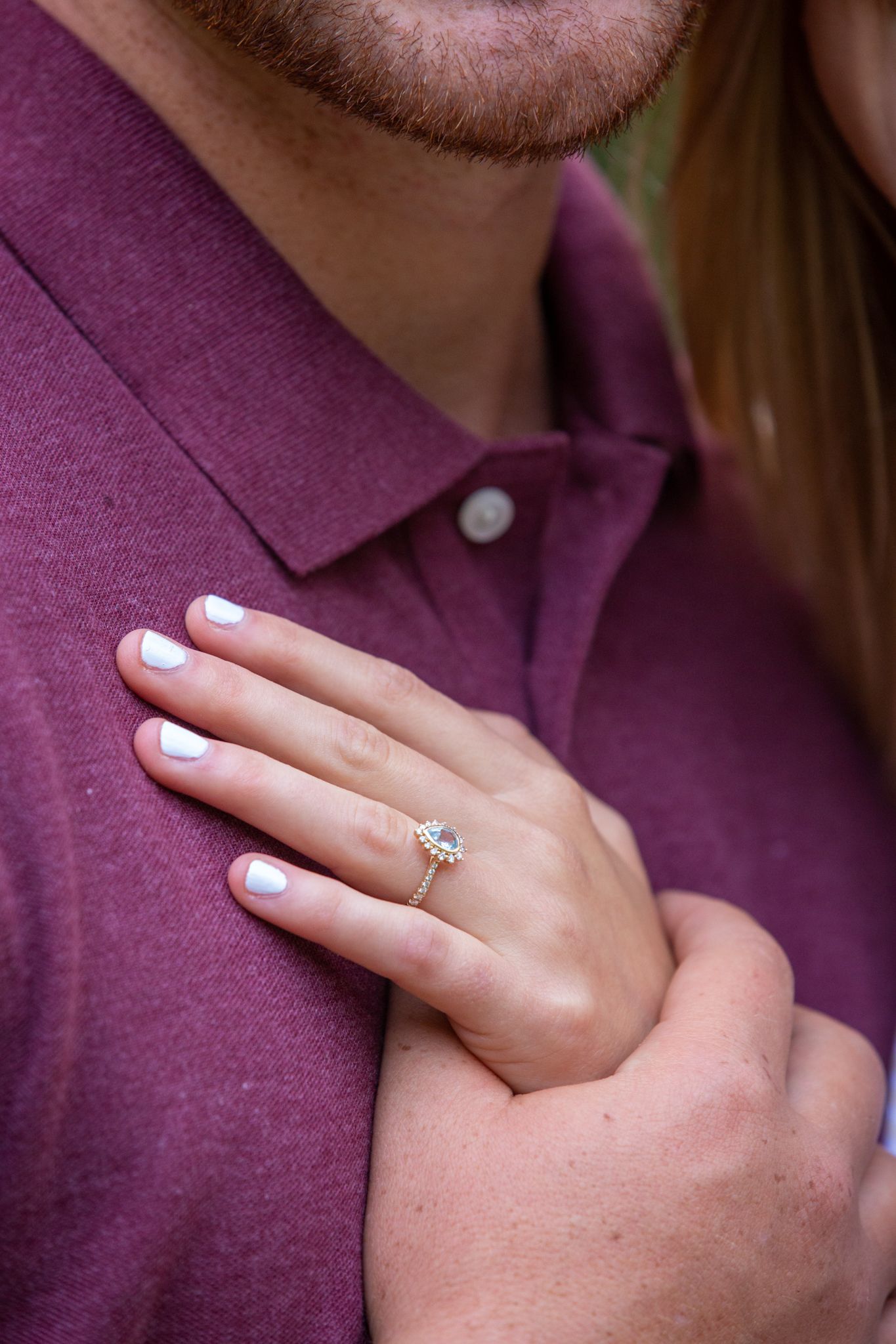 A close-up view of a hand wearing an oval diamond engagement ring resting on someone’s shoulder in a purple shirt.