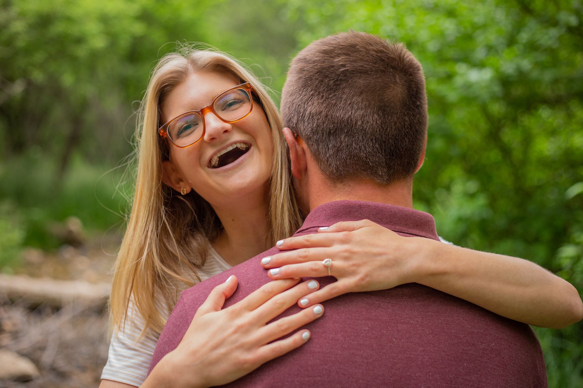 A smiling person wearing glasses hugs another person from behind in a leafy, green outdoor setting.