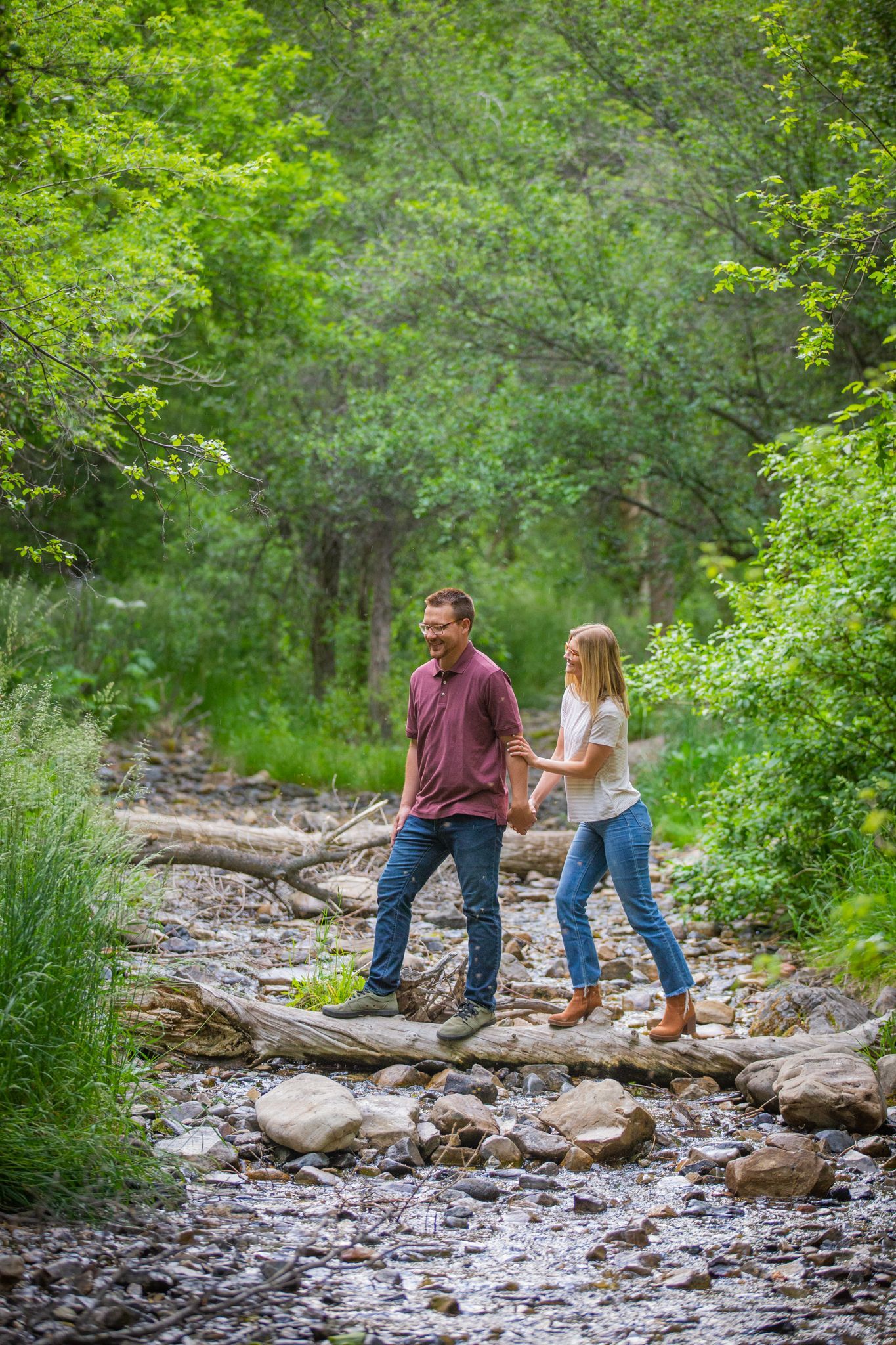 Two people holding hands while carefully walking across a fallen log over a rocky creek in a lush, green forest.