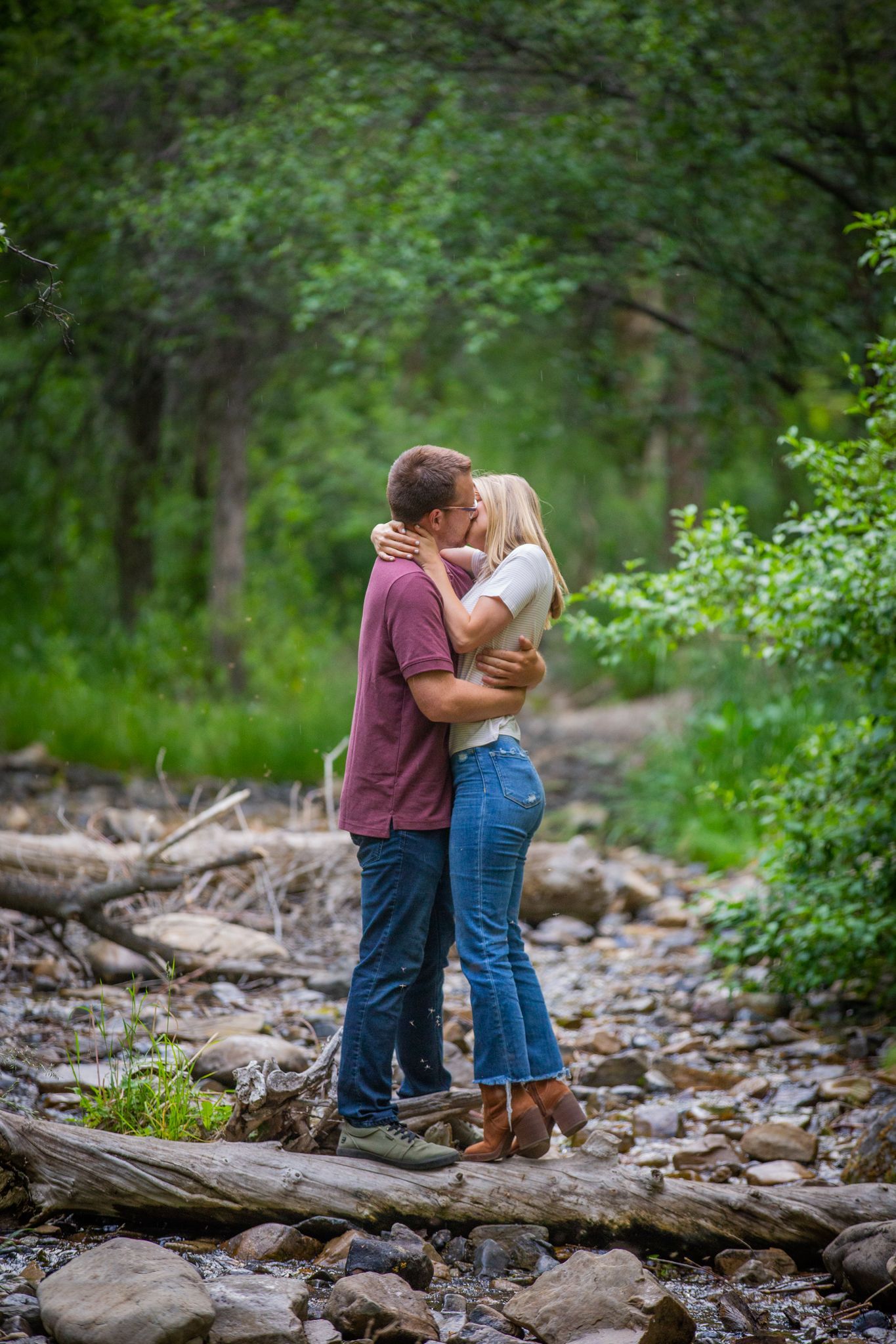 A couple embracing and kissing in a wooded area with a rocky, dry creek bed.