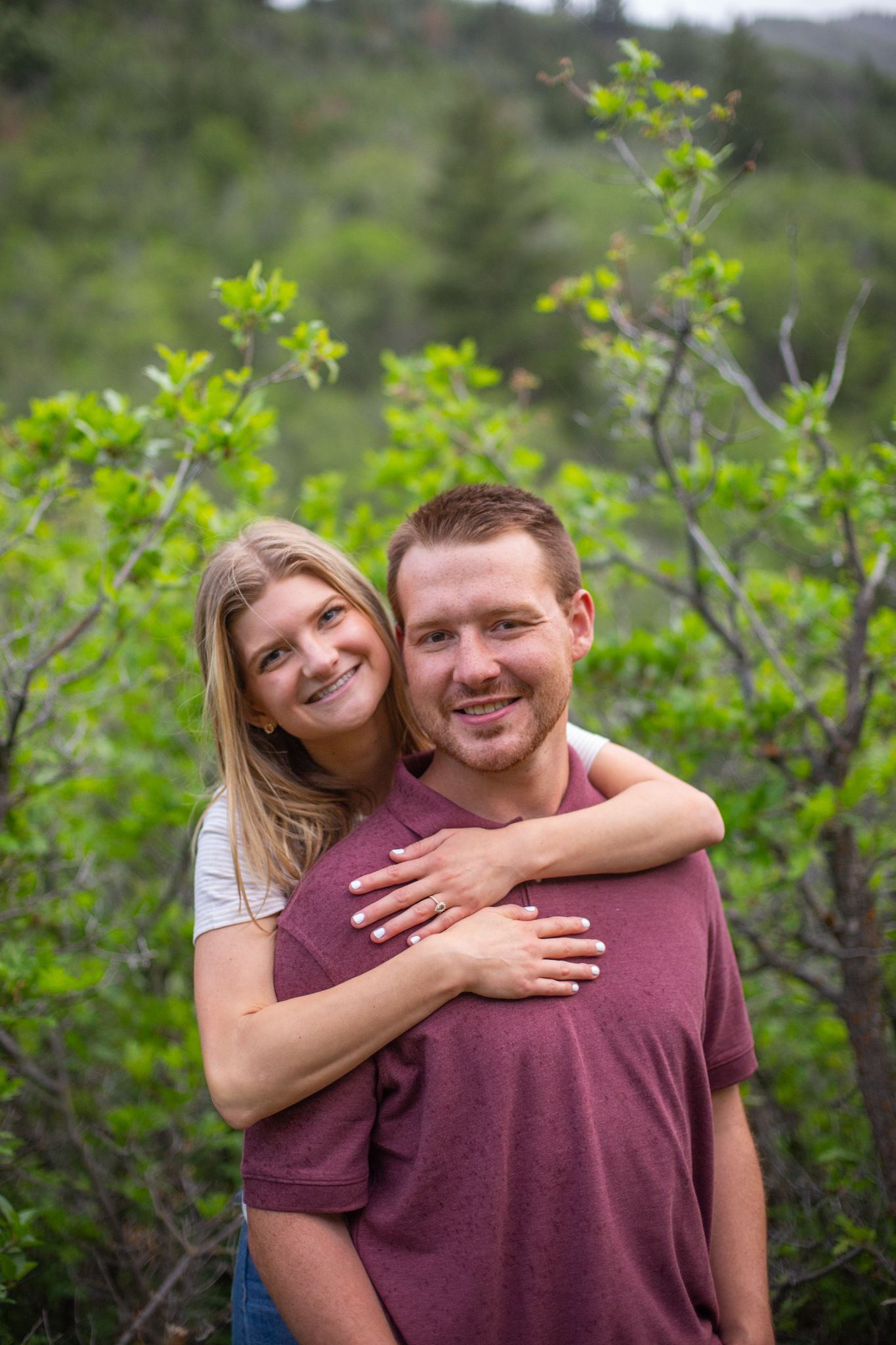 A smiling couple poses outdoors in front of green foliage, with the woman embracing the man from behind.