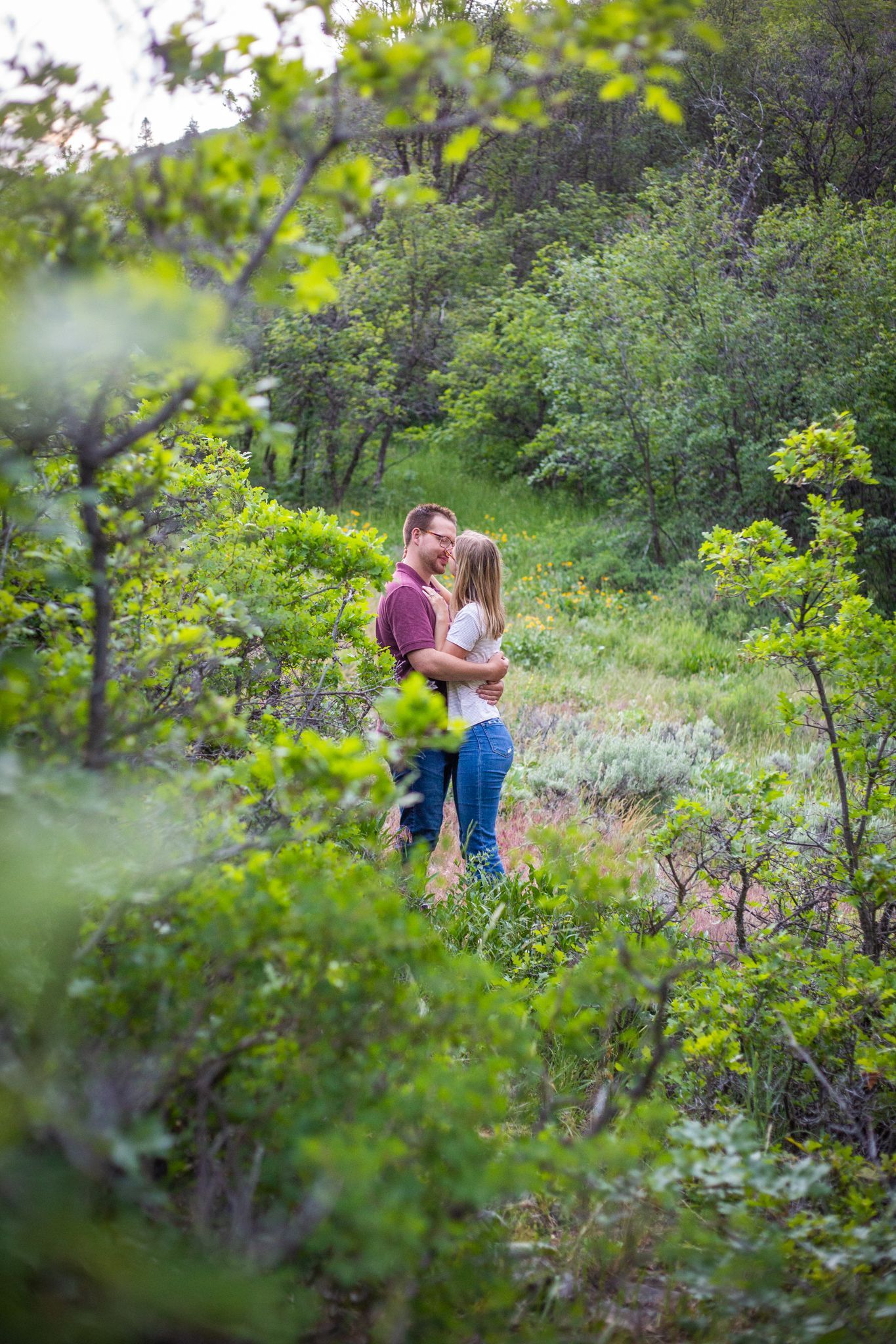 A couple embraces on a dirt trail surrounded by lush green foliage and trees in a forest.