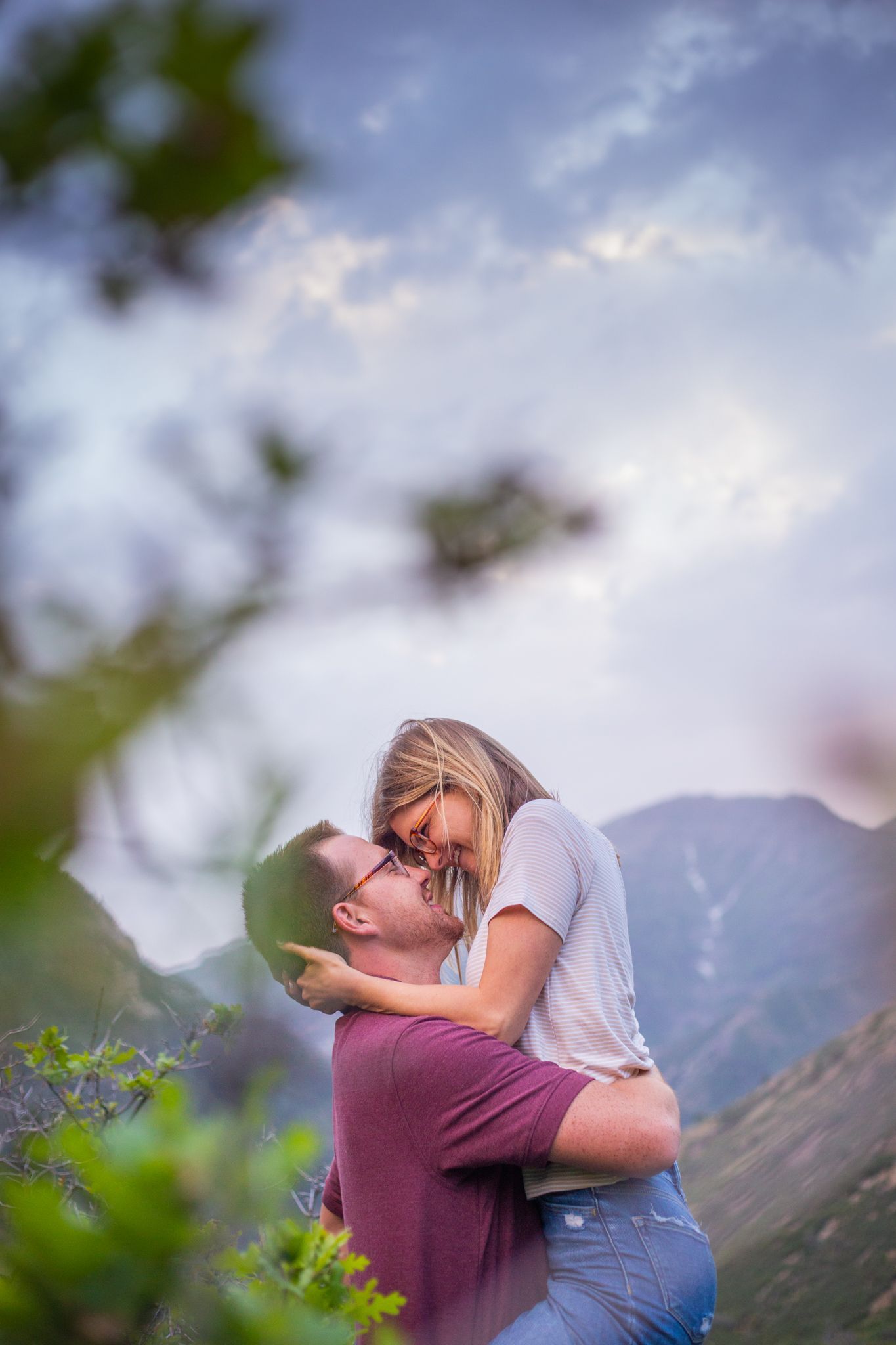 A couple embraces affectionately outdoors with a mountain landscape and cloudy sky in the background.