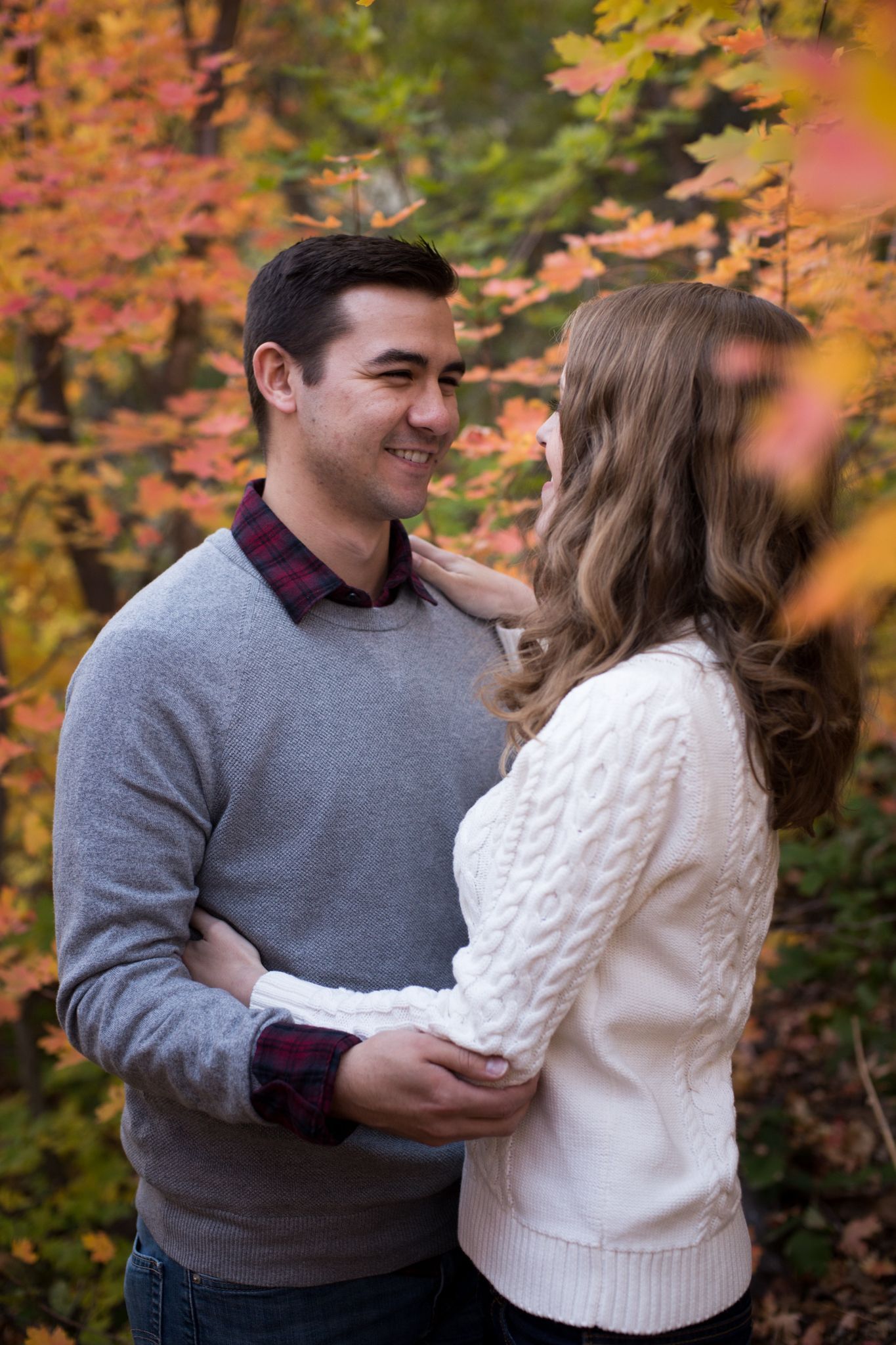 A couple standing close and smiling at each other in a wooded area surrounded by autumn leaves.