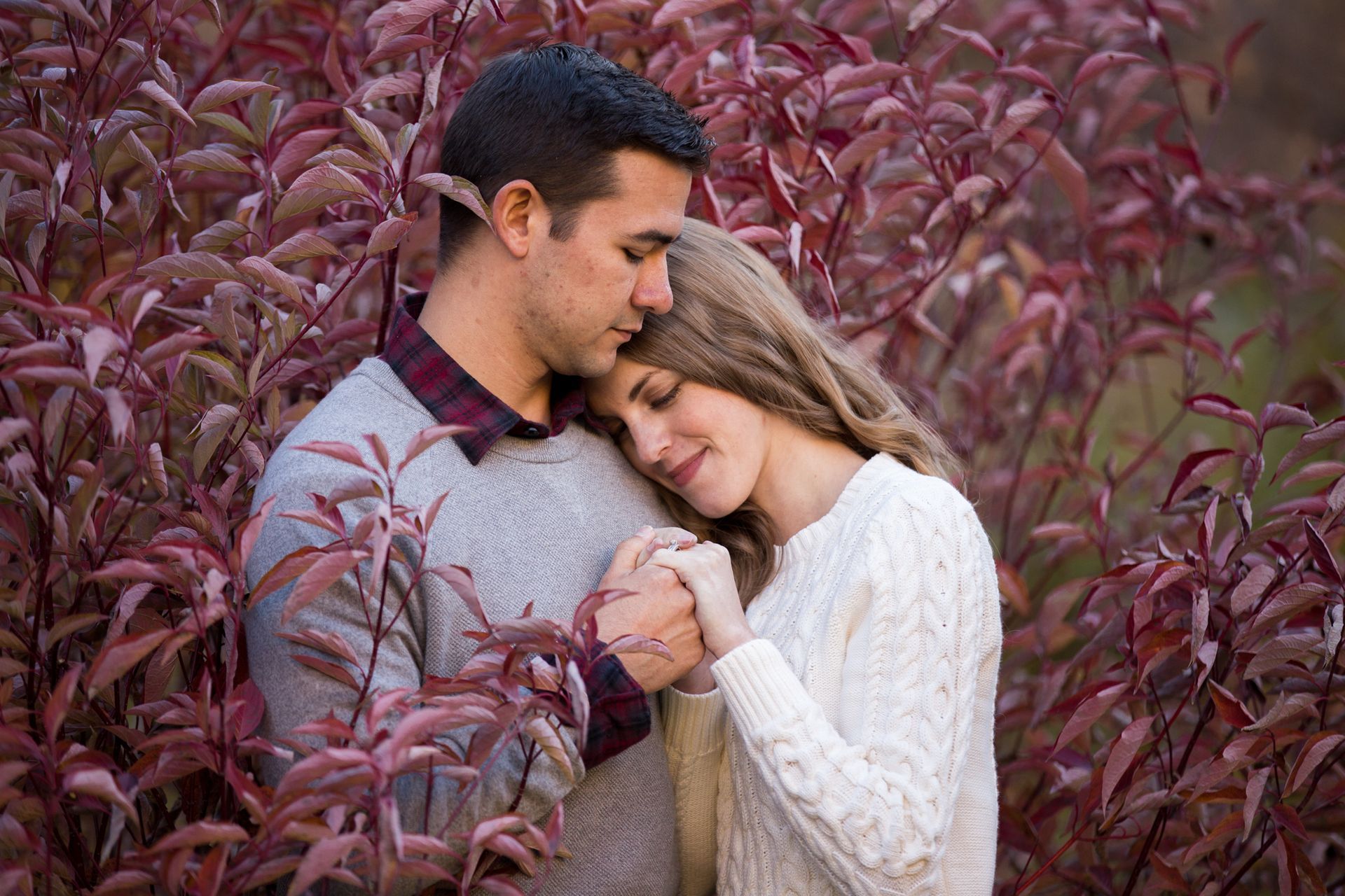 A couple stands close together, holding hands with their heads touching amidst a background of deep red autumn foliage.