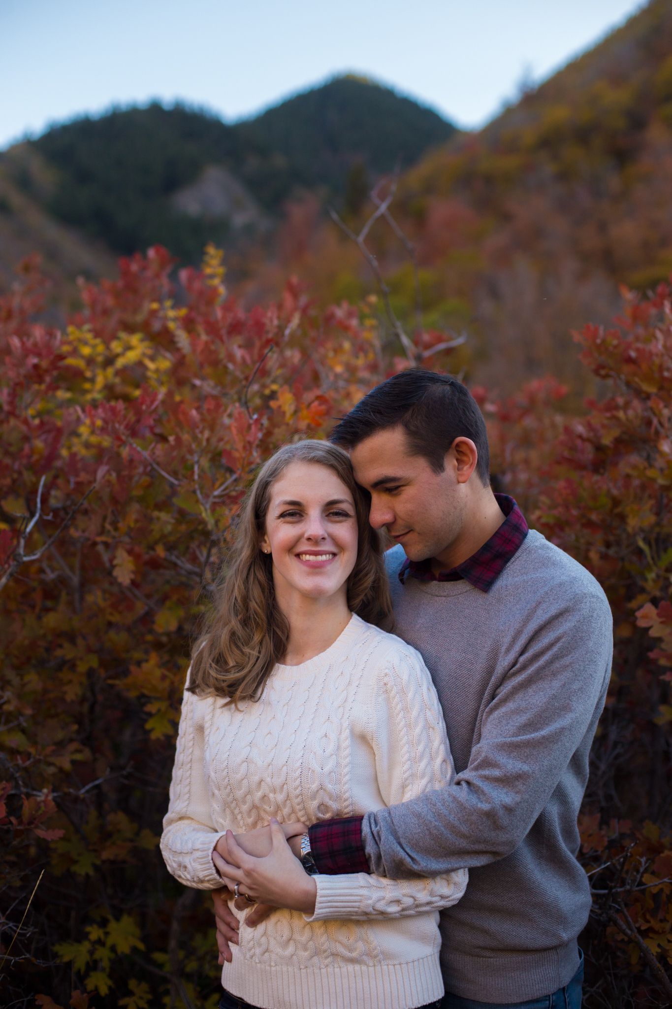 A couple embraces in front of autumn foliage with mountains in the background.