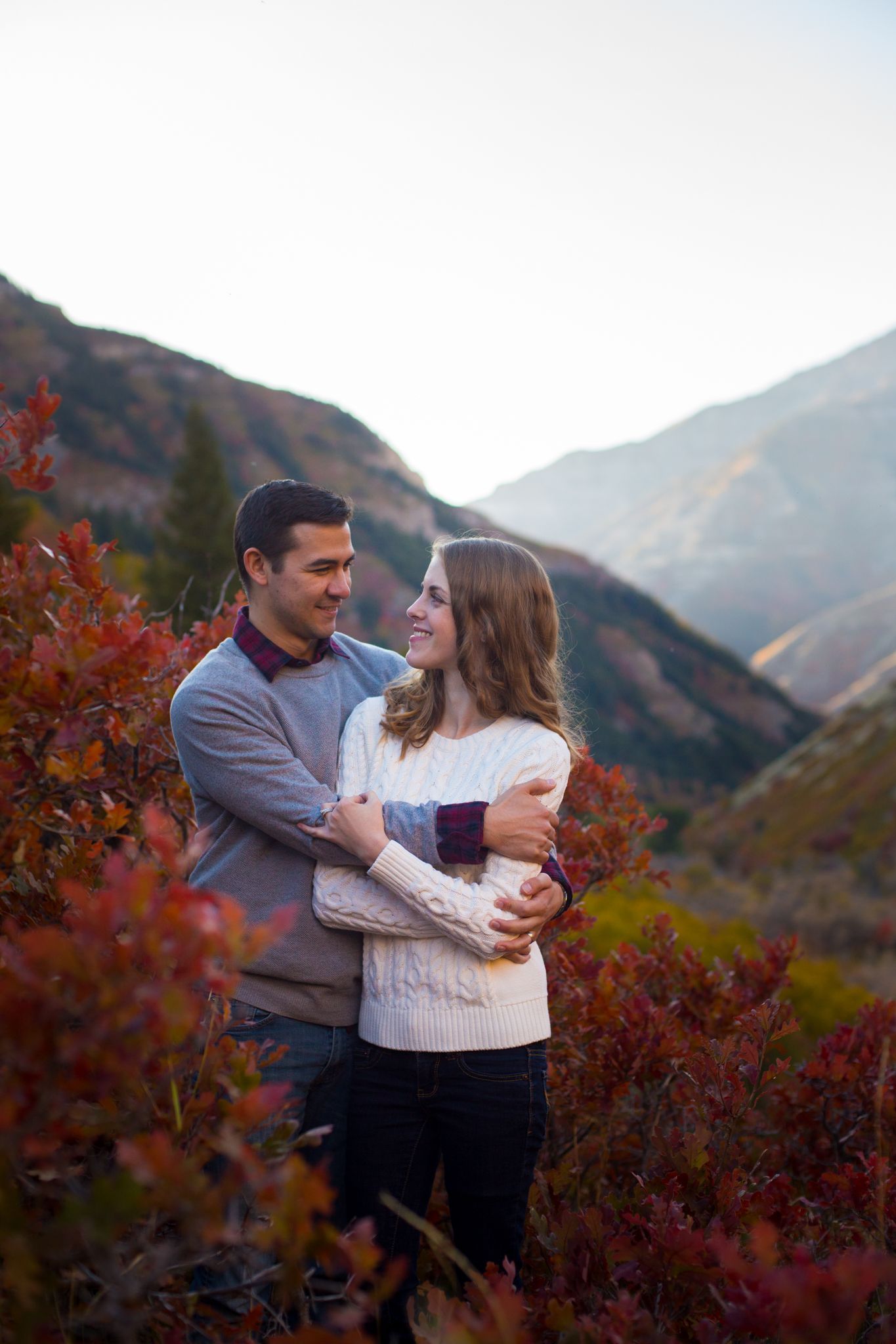 A couple embraces in a mountainous landscape filled with vibrant red and orange autumn foliage.