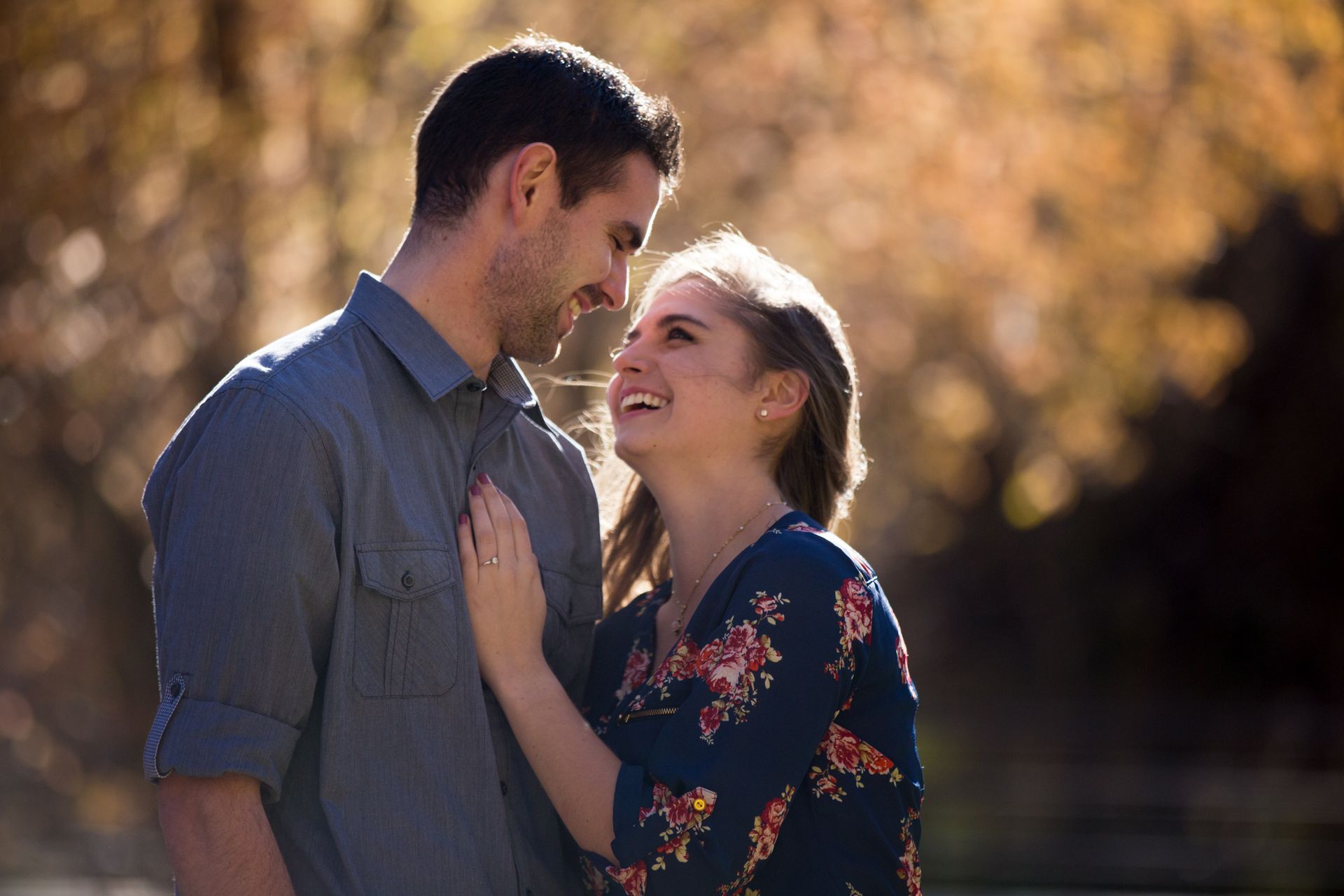 A couple in casual clothing looks at each other and smiles affectionately in a softly lit, golden-toned outdoor setting.