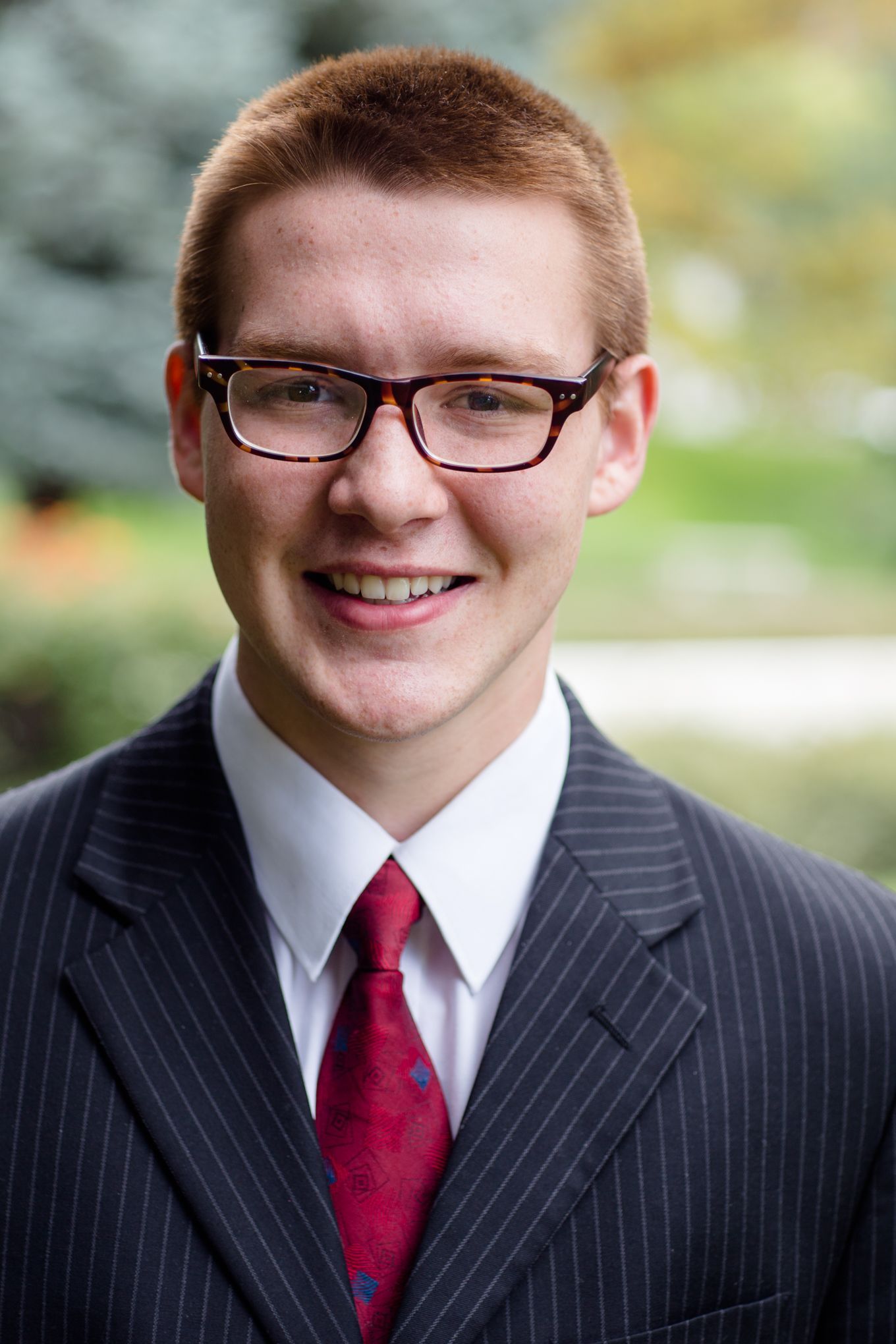 A person in a pinstripe suit, white shirt, and red tie with glasses smiling warmly against an outdoor, blurred background.
