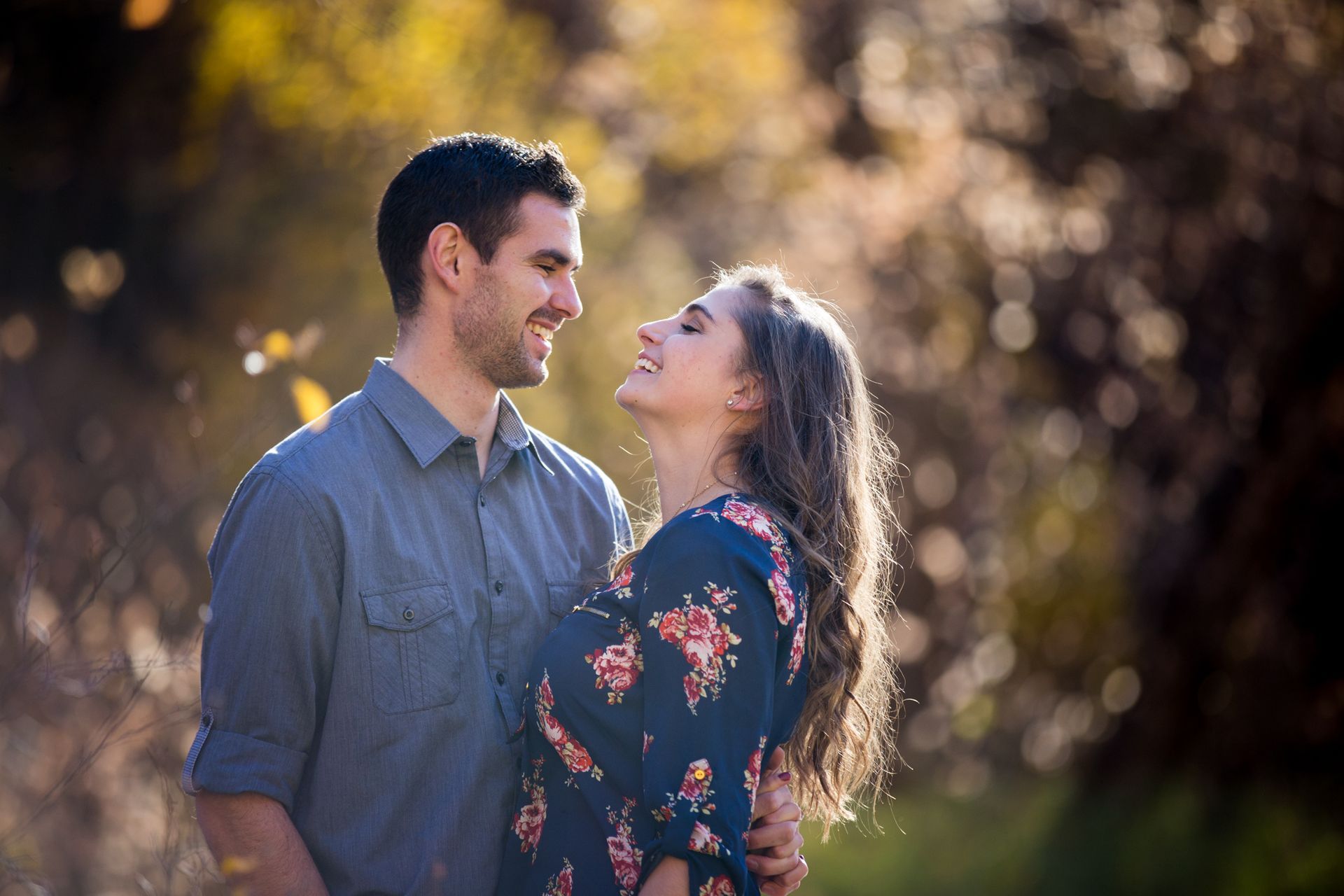 A man in a gray shirt and a woman in a floral dress look at each other and smile in a sunlit, out-of-focus park setting.