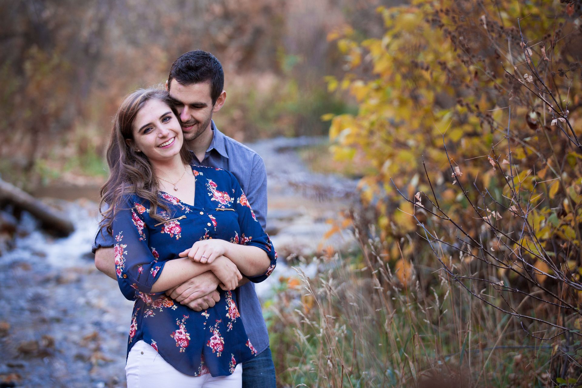 A couple stands smiling in an embrace by a stream, surrounded by fall foliage.
