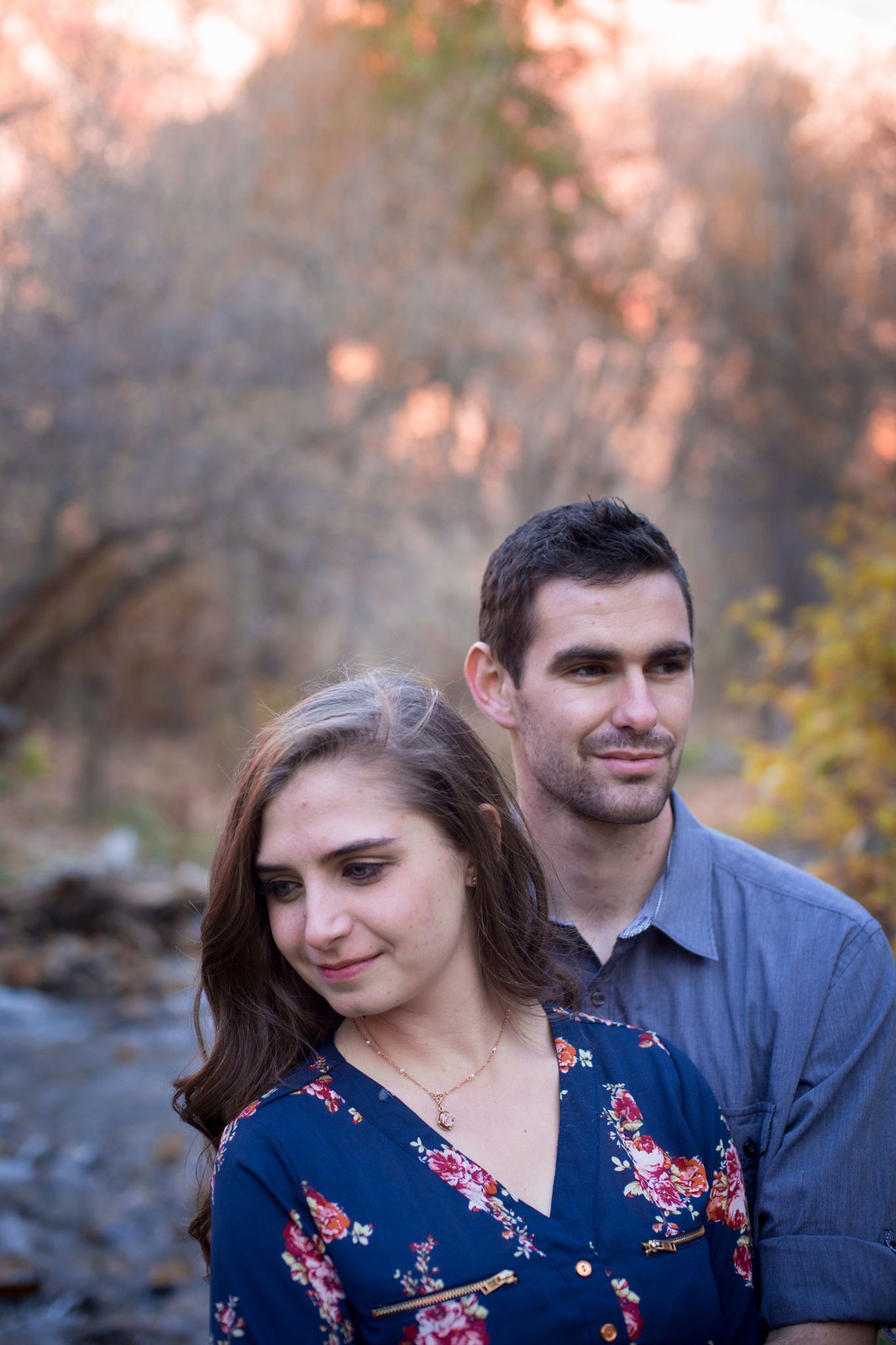 A man stands behind a woman with wavy hair in a floral dress, both posing outdoors in a softly lit, natural setting.