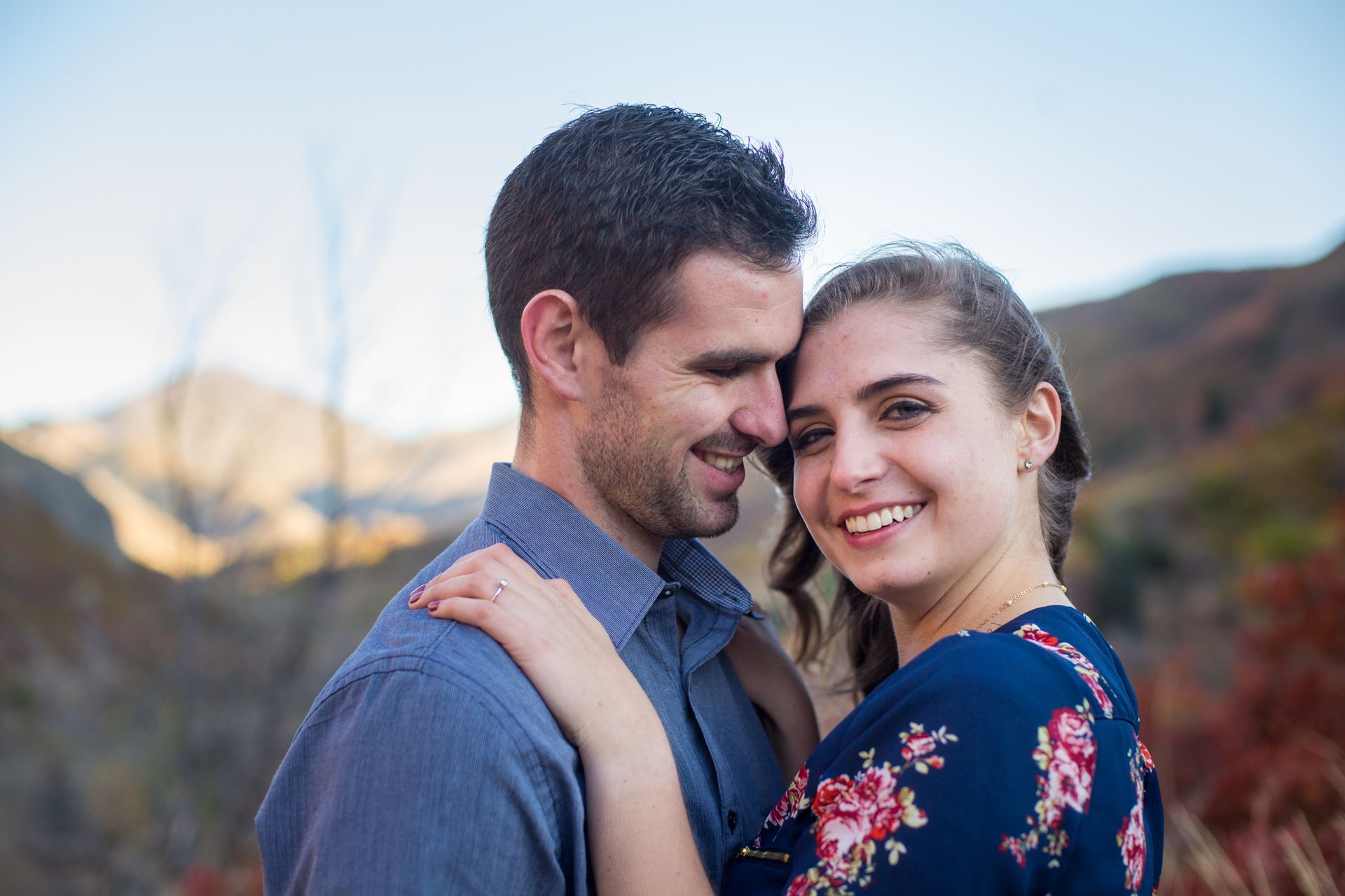 A couple smiling closely together outdoors with mountains in the background during golden hour.