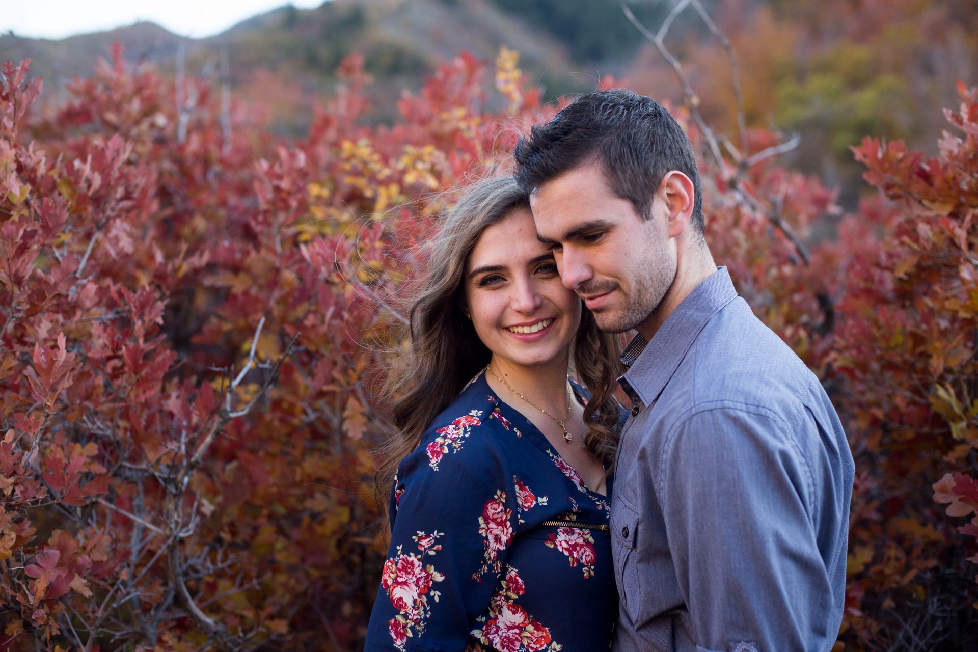 A couple smiles together in front of autumn foliage with reddish-orange leaves.