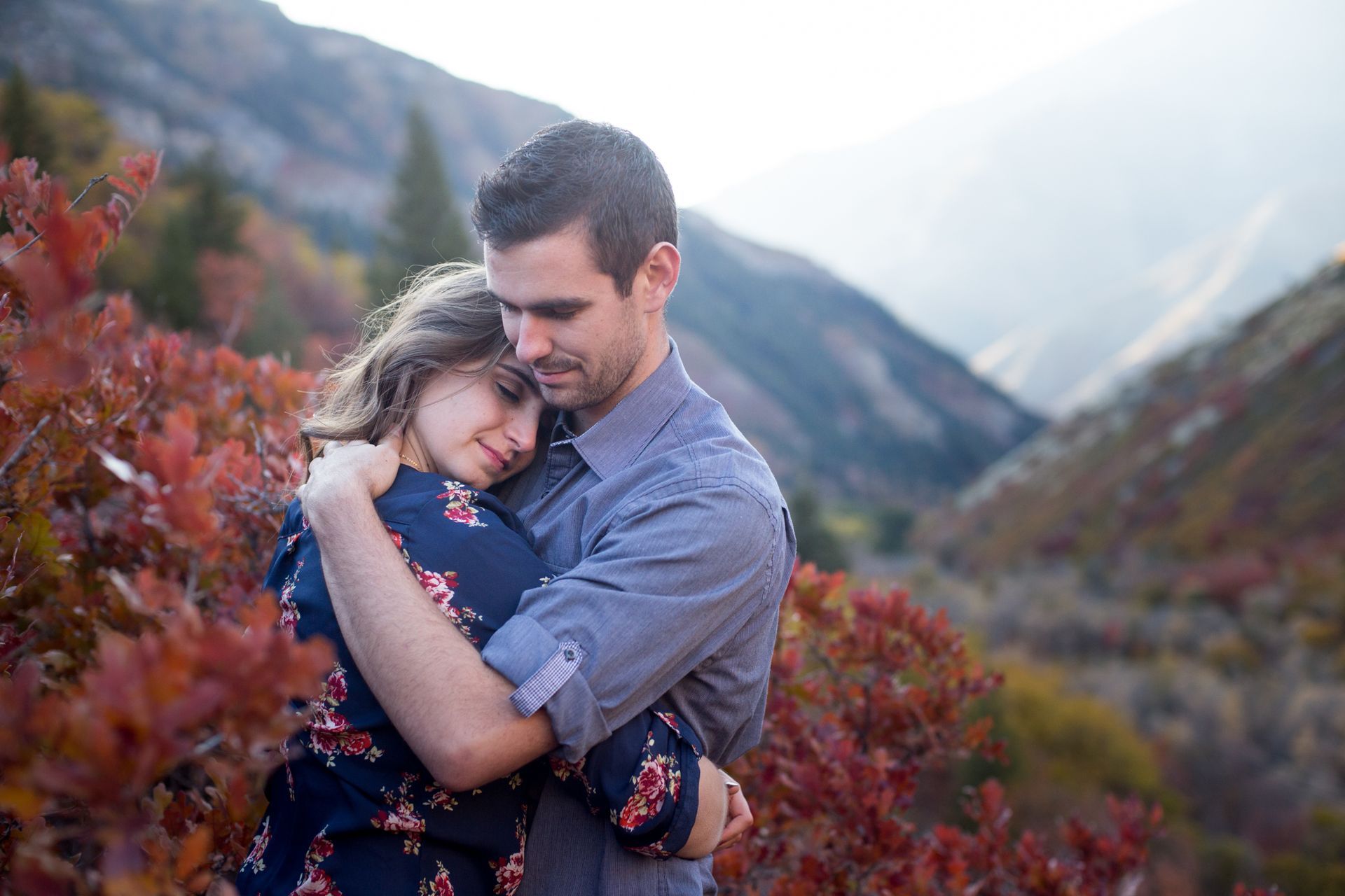 A couple embraces warmly while standing among autumn foliage with mountains in the background.