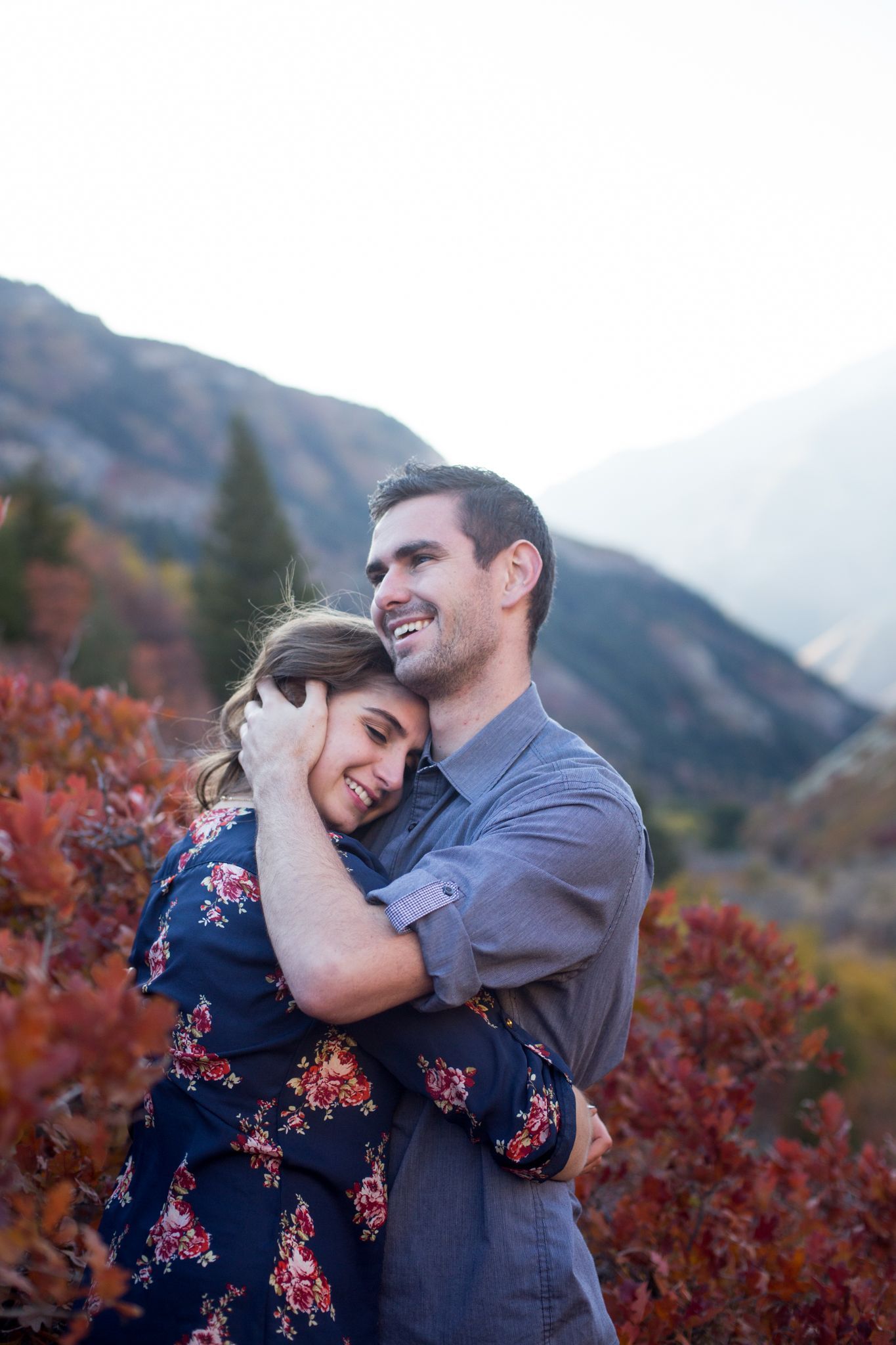 A couple embraces outdoors among fall foliage with a mountain background.