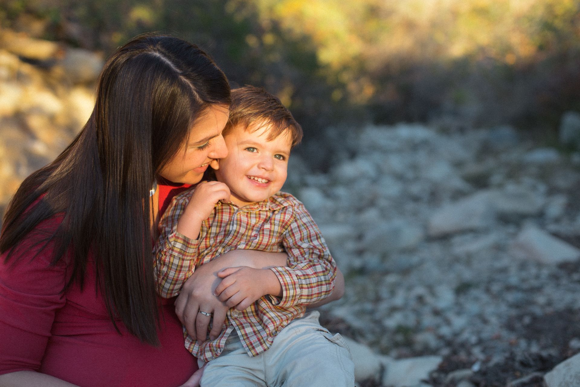 A woman in a red shirt embraces a smiling child in a plaid shirt outdoors at golden hour.