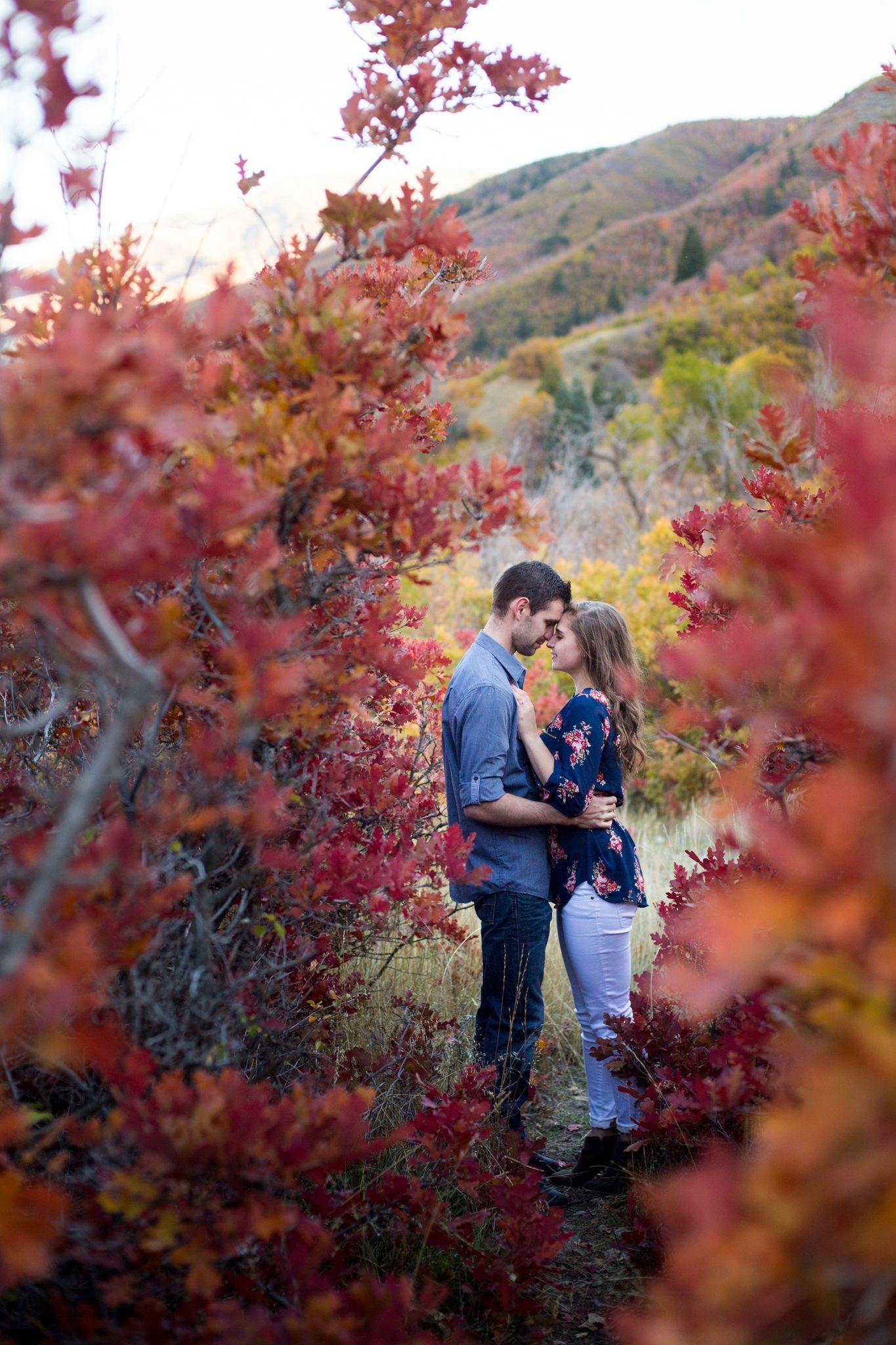 A couple embraces in a forest during autumn, framed by vibrant red and orange foliage with mountains in the background.