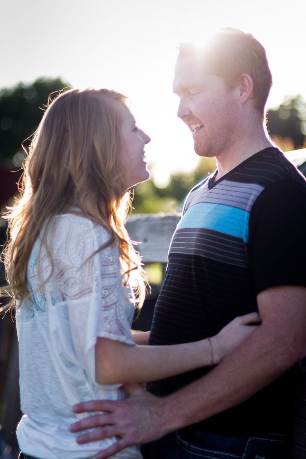 A couple stands face-to-face outdoors in backlighting, smiling at each other with their arms around one another.