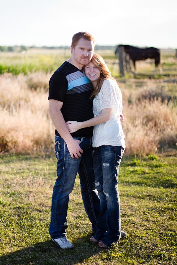 A couple embraces in a sunlit field, with a horse grazing in the background.