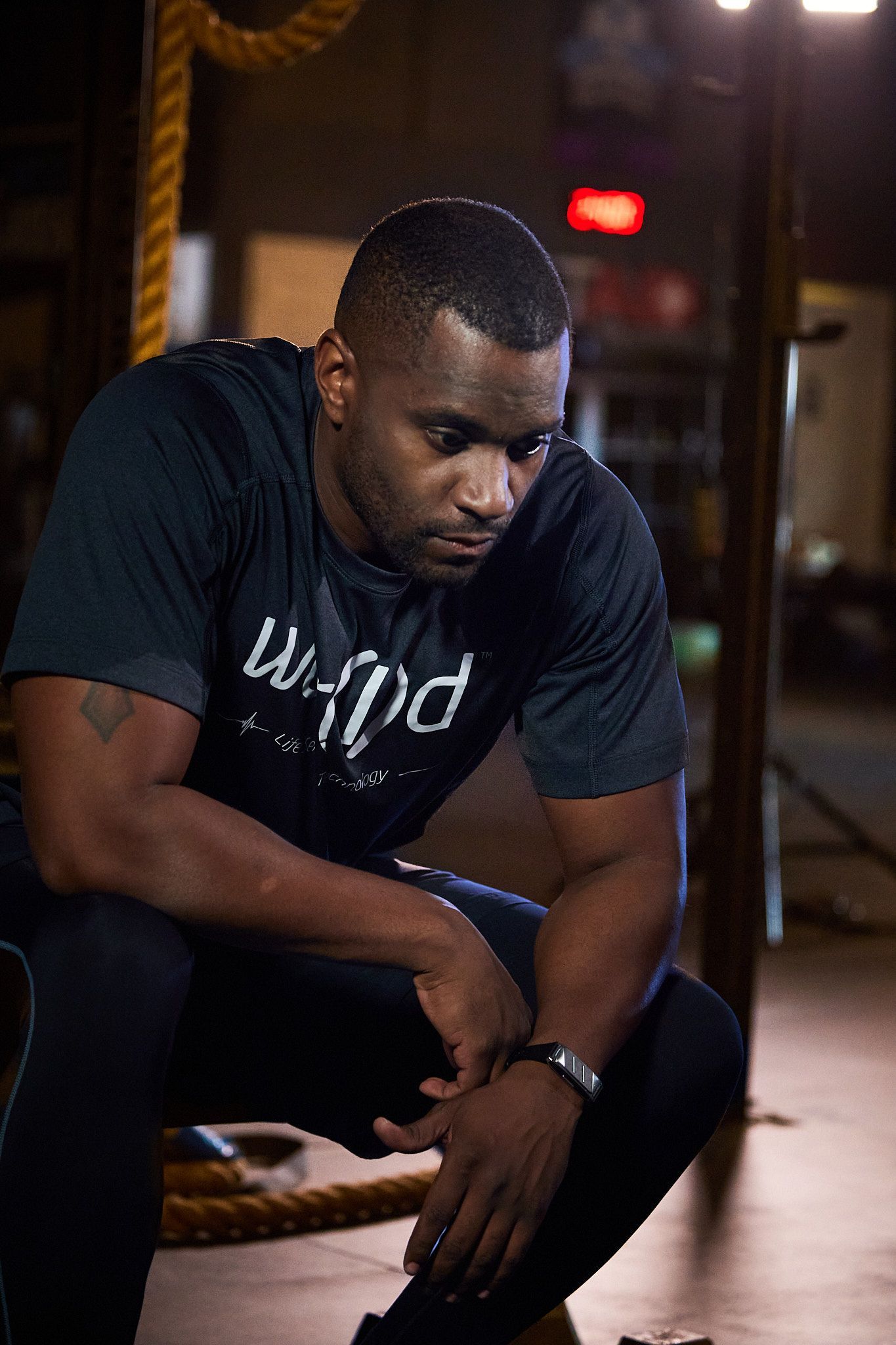 A person in athletic gear sits in a gym, looking downward with a focused expression, leaning forward with hands on knees.
