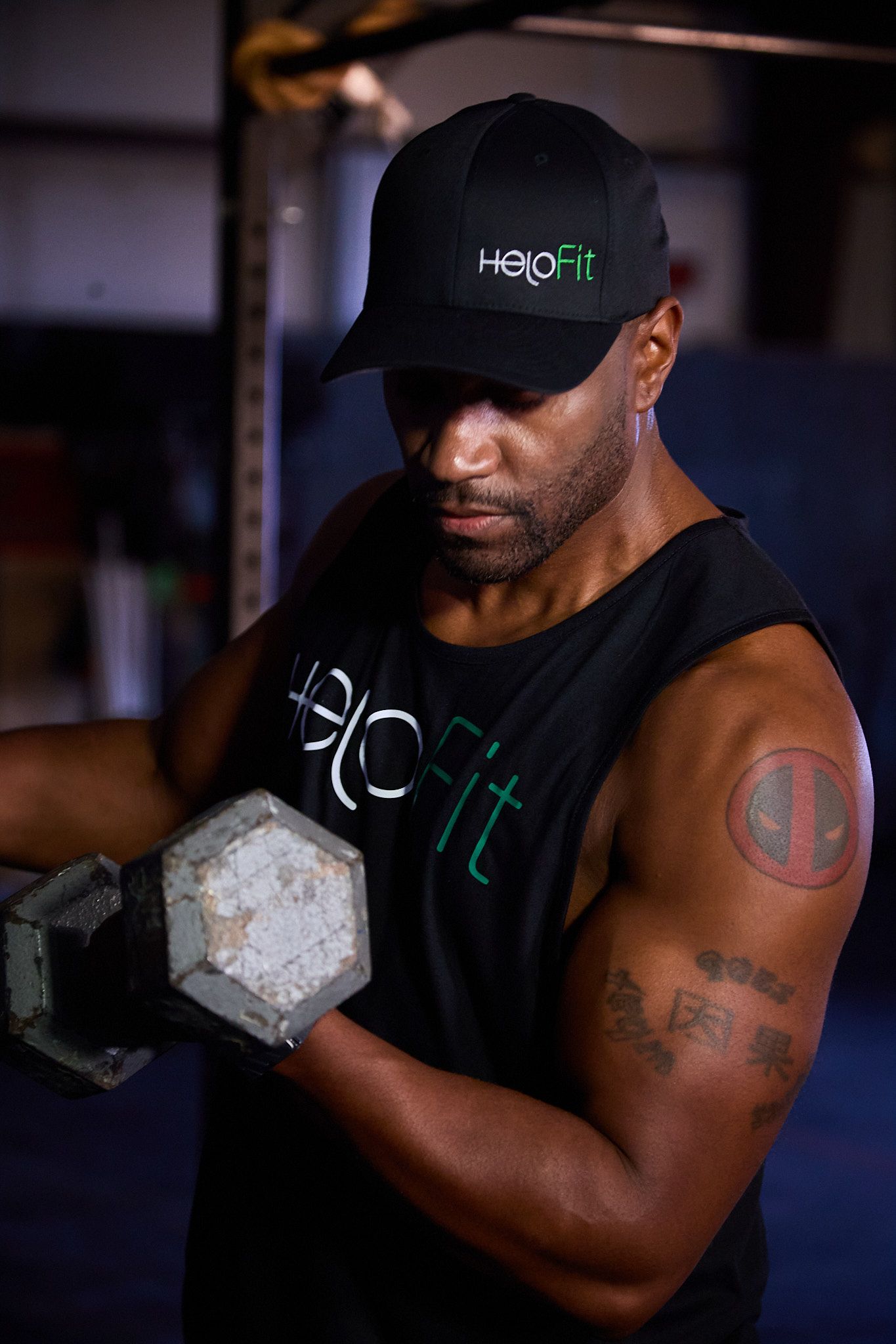 A person wearing a black tank top and cap performs a dumbbell curl in a gym, revealing a tattoo on their arm.