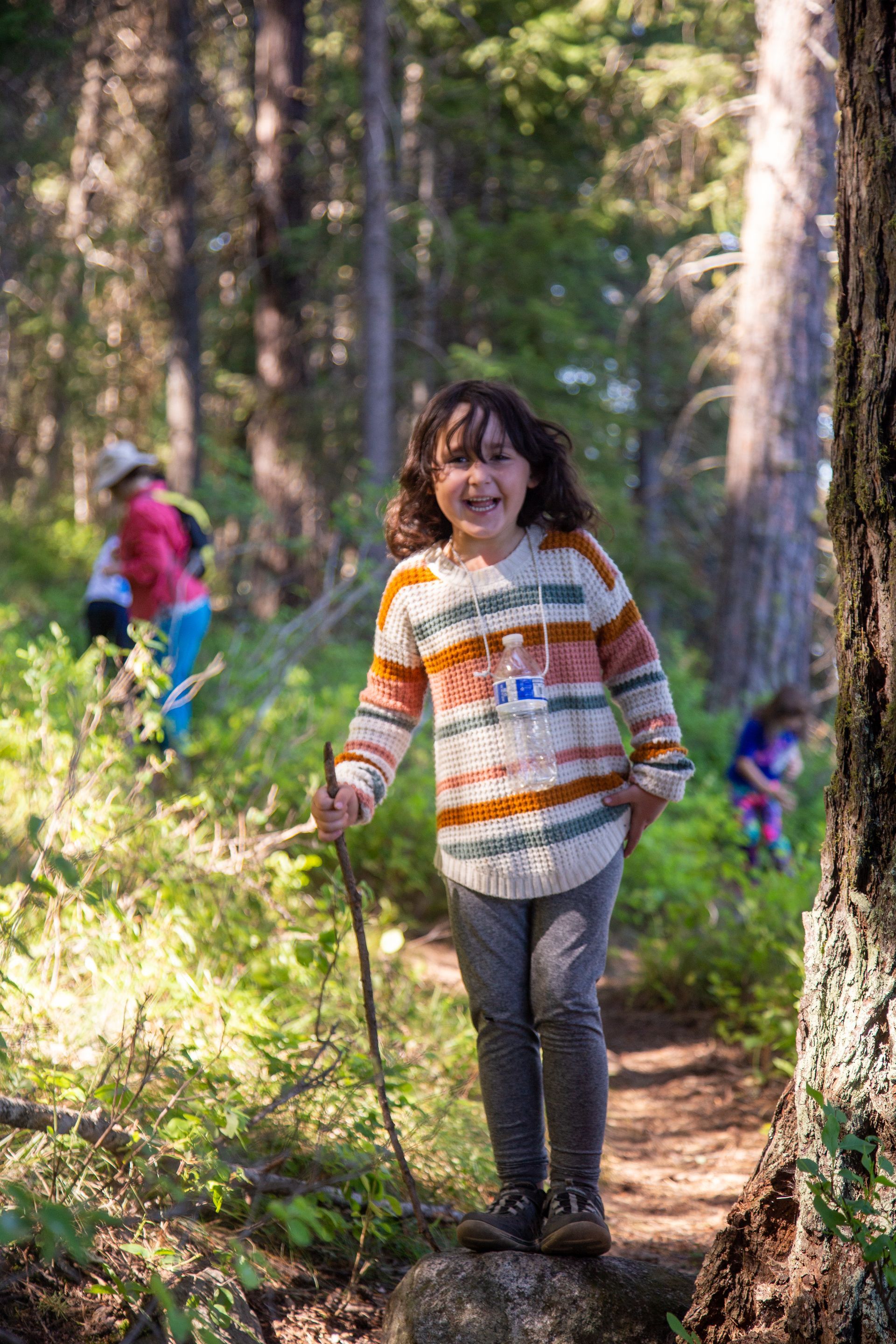 A smiling child in a striped sweater stands on a trail in a forest, holding a walking stick.