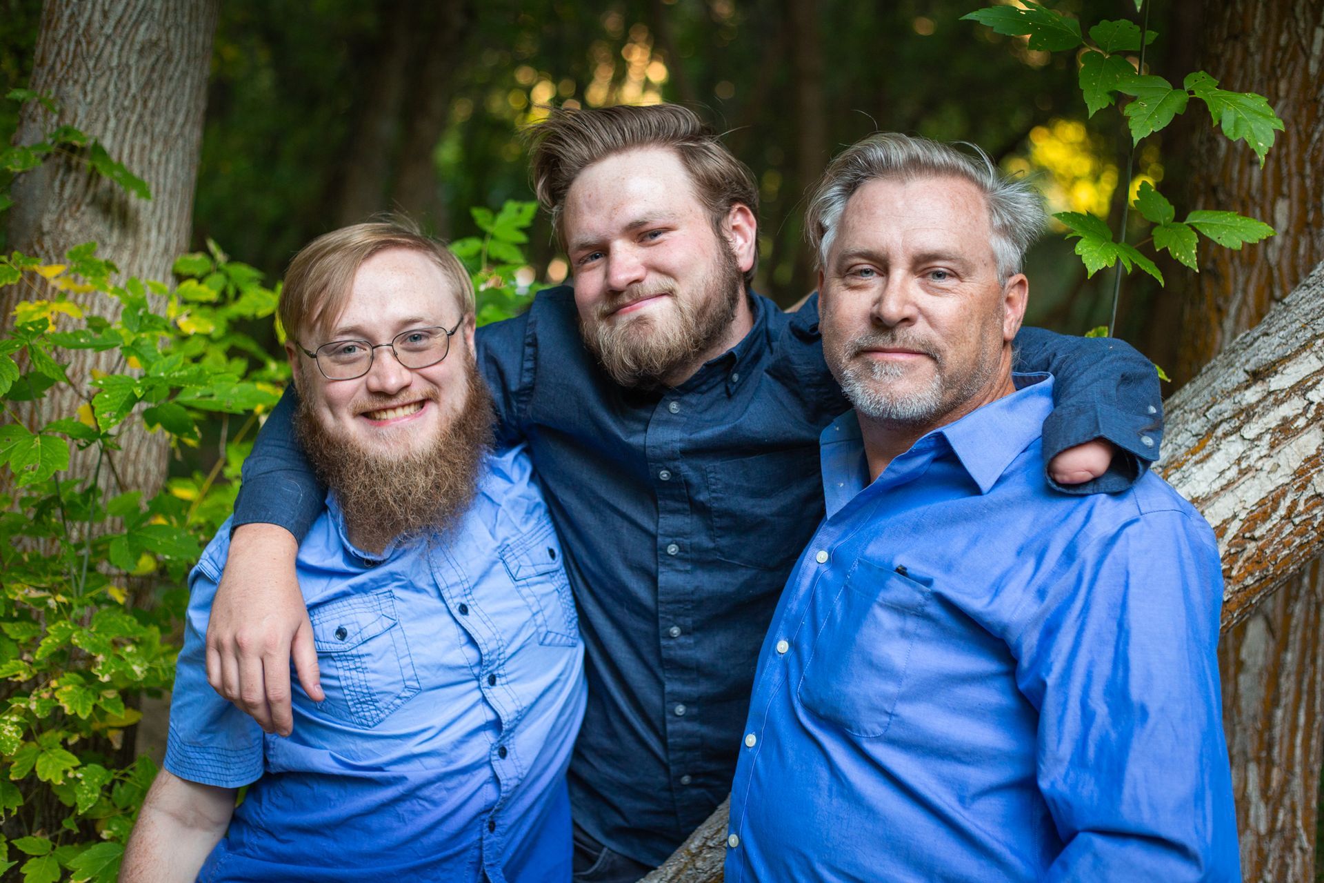 Three people with arms around each other, wearing blue button-down shirts, posing together in a wooded area.
