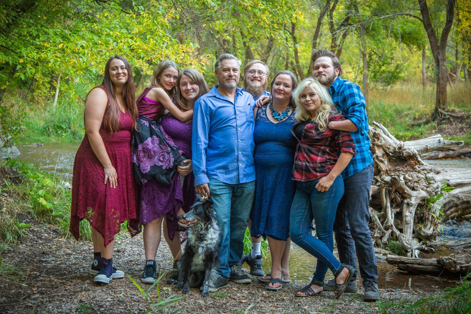A group of nine people and a dog stand smiling together in a wooded, outdoor setting near a fallen tree.