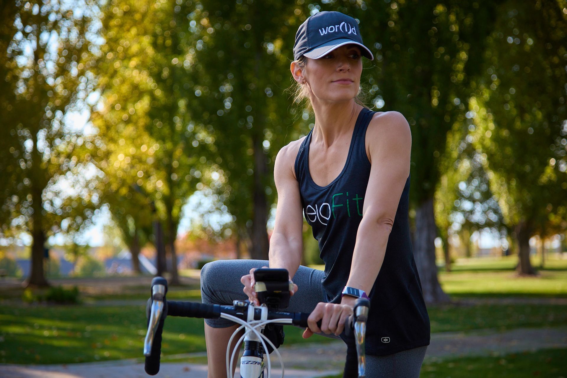 A person wearing a cap and a dark tank top stands outdoors with a bicycle, holding a phone on the handlebars.